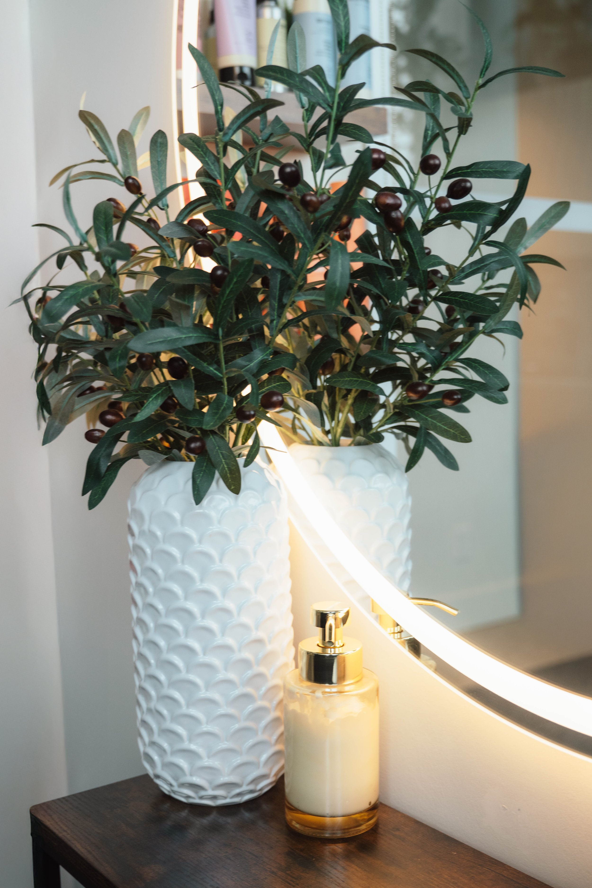 A decorative scene with a large white textured vase holding a plant with green leaves and dark berries on a dark wooden surface. There's a candle dispenser with lotion or soap in front of a backlit mirror.