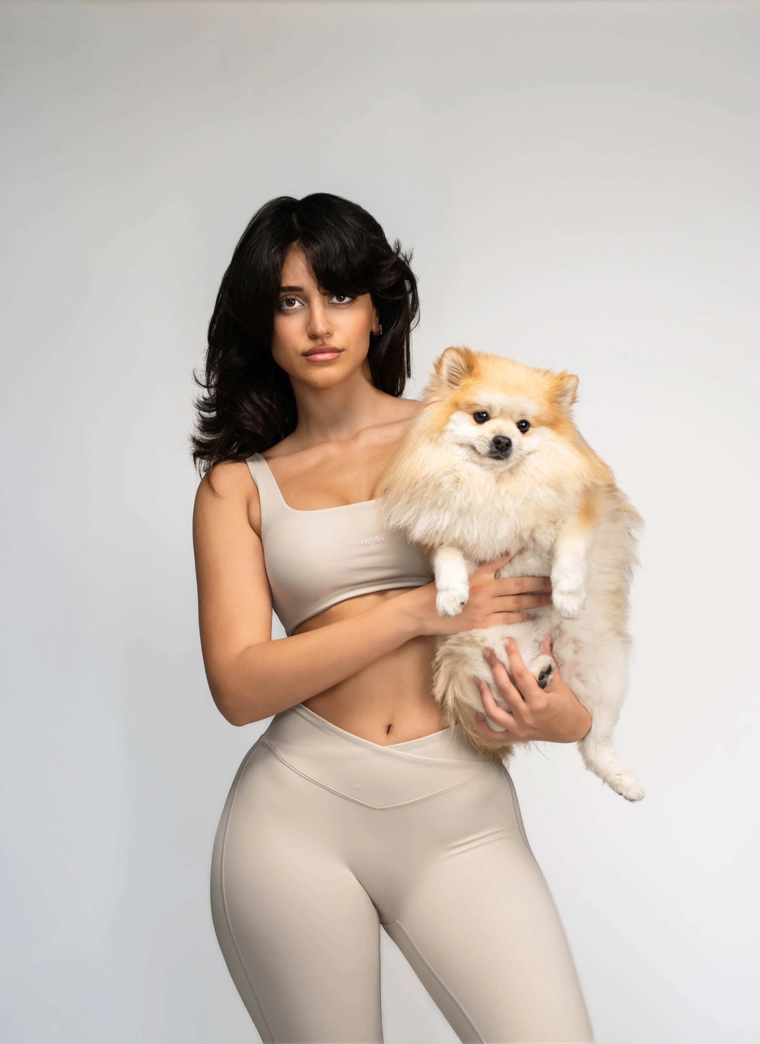 A woman with dark hair wearing a beige sports bra and matching leggings, holding a small fluffy dog with tan and white fur against a plain background.