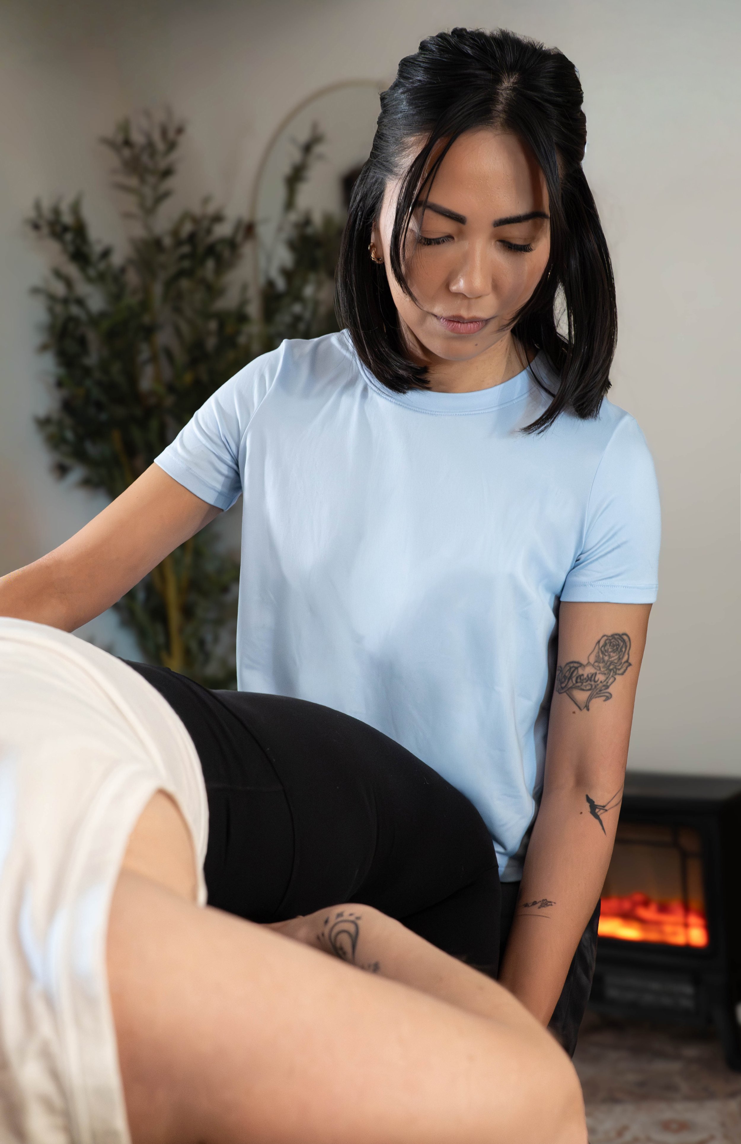 Woman in light blue shirt providing massage in room with potted plant and electric fireplace.