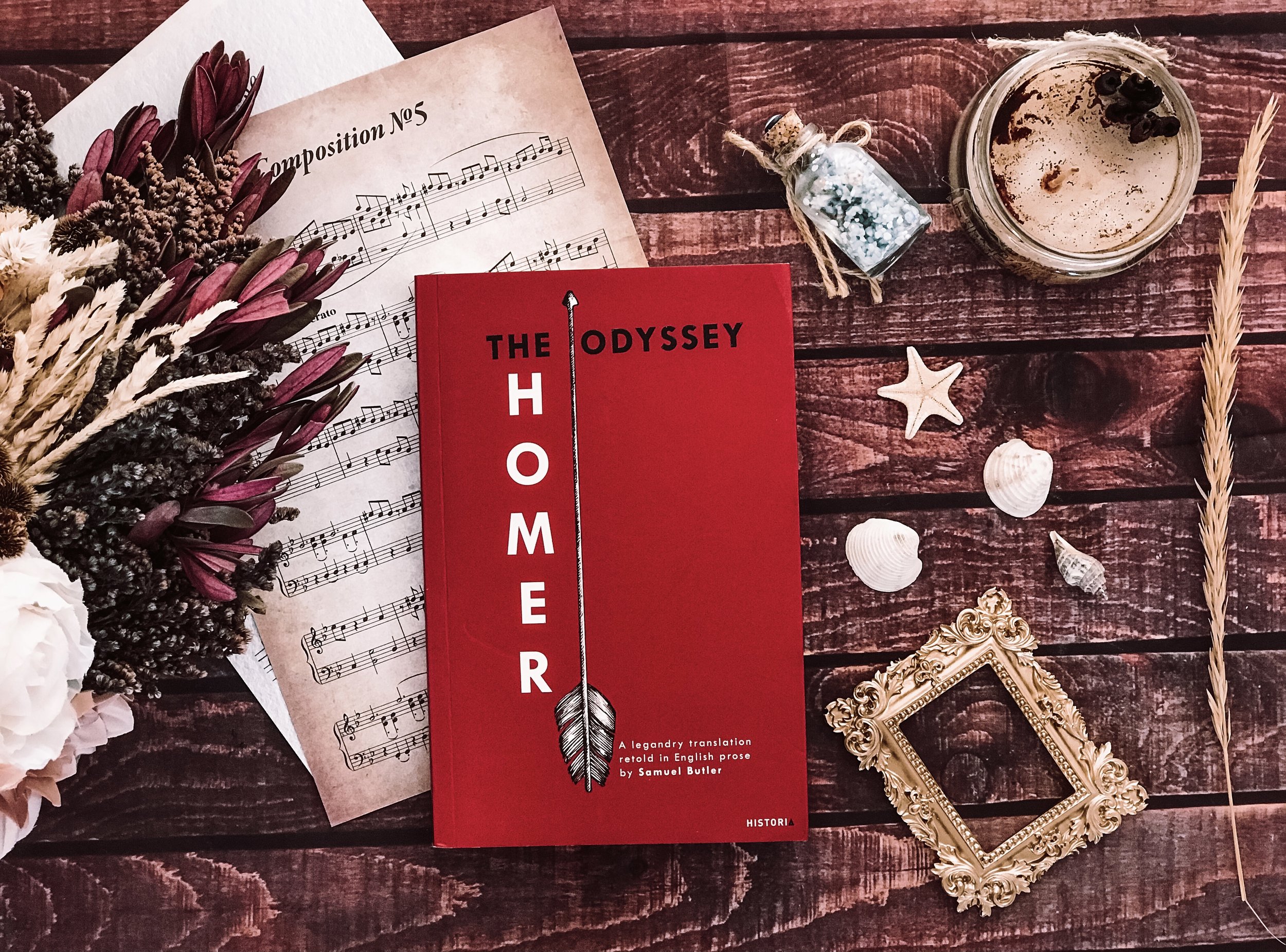 A red book titled 'The Odyssey Homer' lies on a dark wooden surface, surrounded by seashells, starfish, a gold ornate picture frame, a glass jar with a candle and pine cones, a small jar of glitter, a candle with black beans, sheet music, and dried f