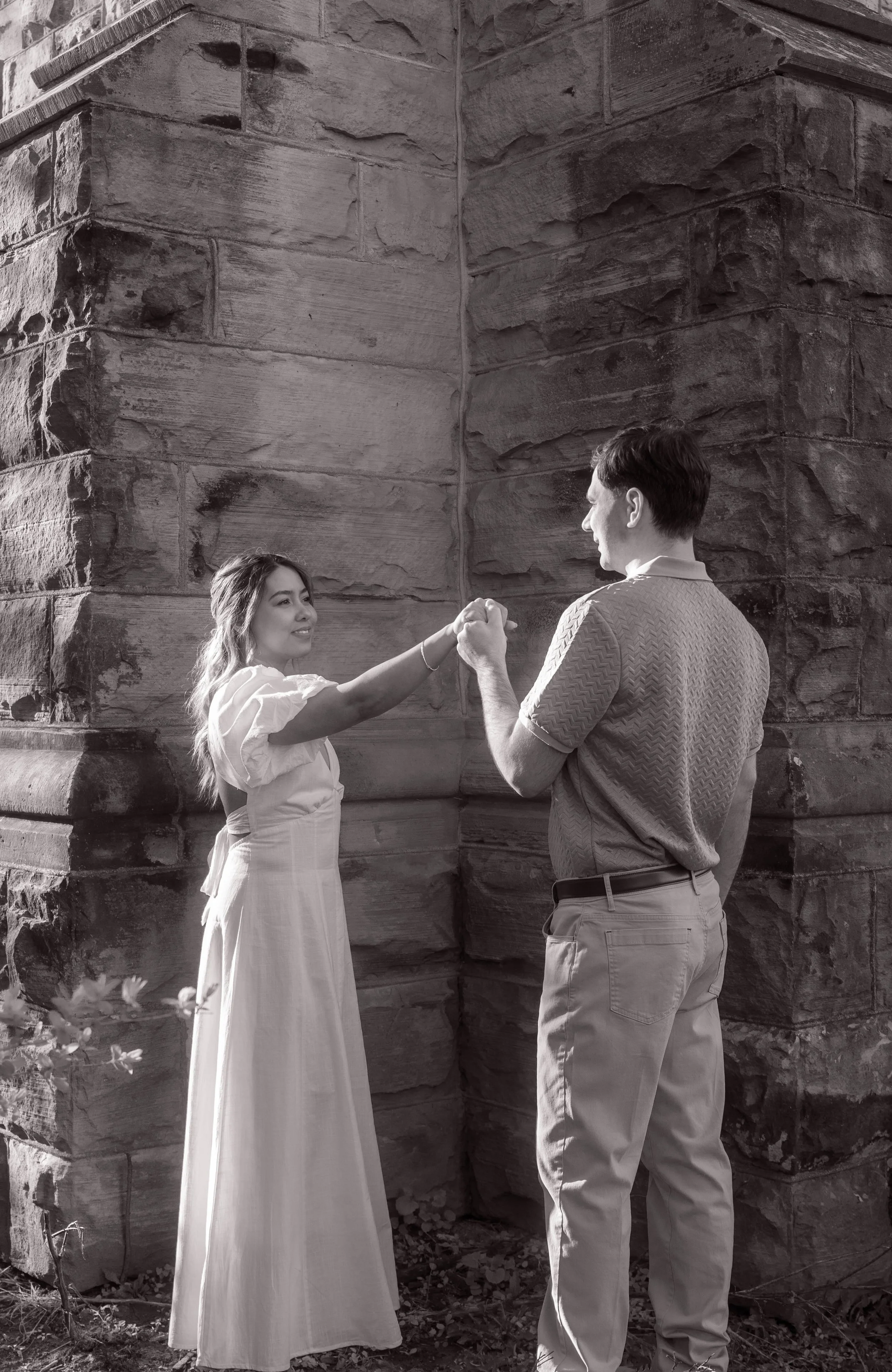 Couple holding hands during a romantic engagement session at Battlefield Park in Hamilton, Ontario, captured in black and white against historic stone architecture