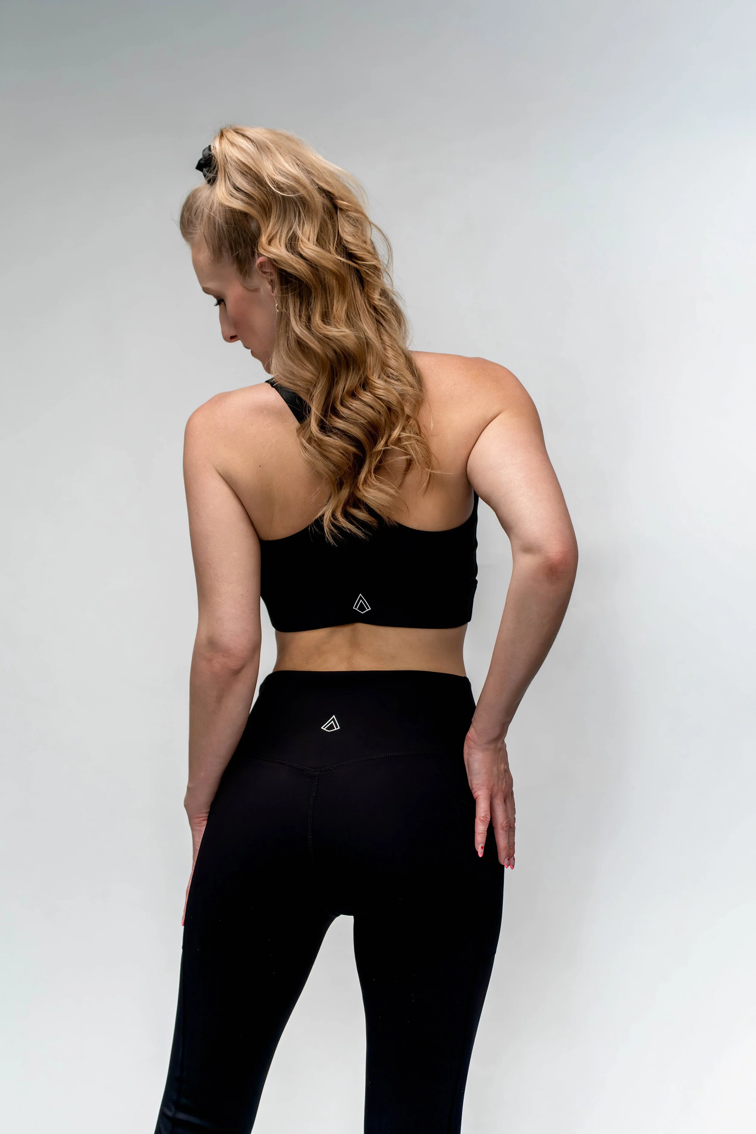 Back view of a woman with long, wavy blonde hair wearing a black sports bra and black leggings against a plain light background.