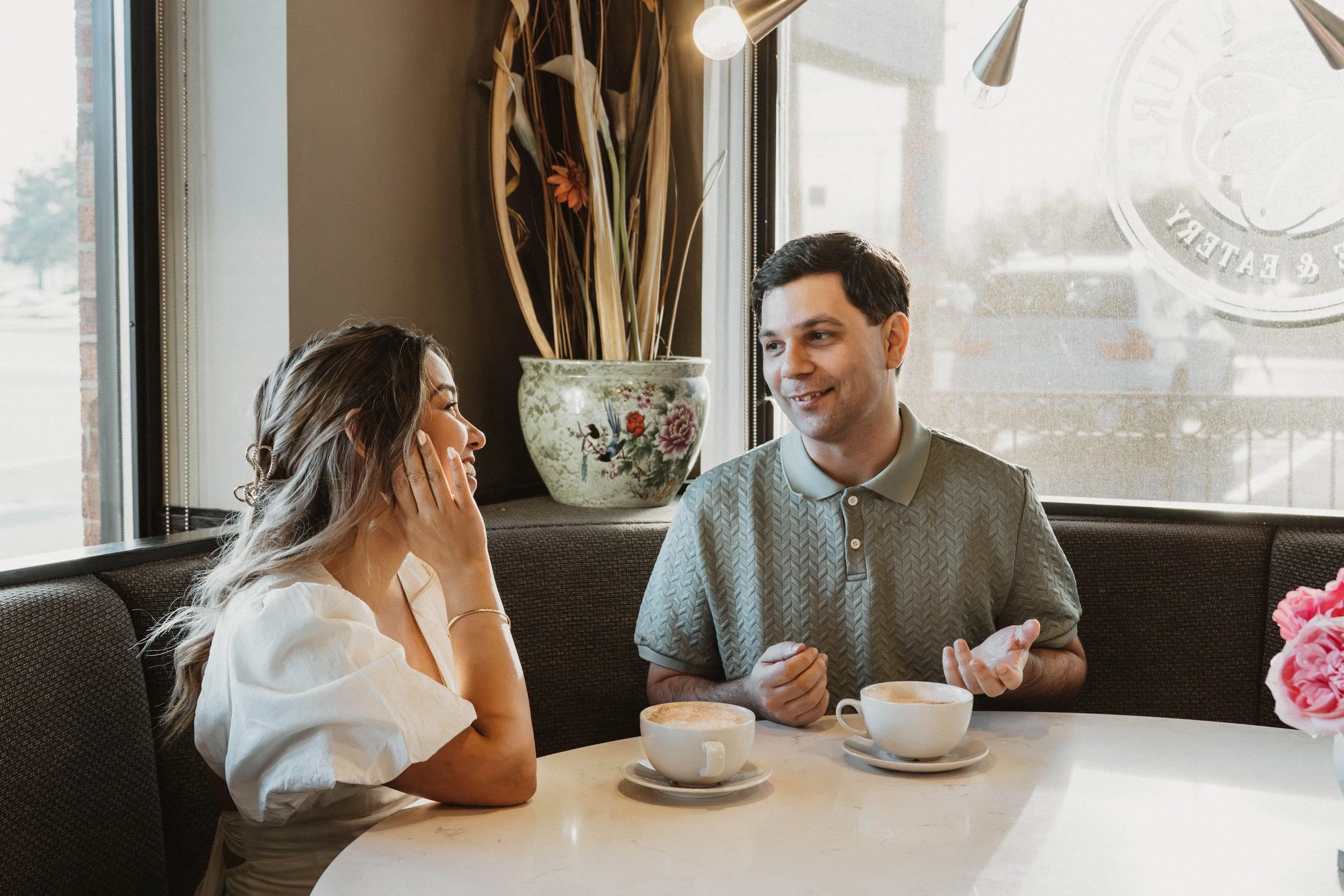 A man and woman sitting in a restaurant near a window, engaged in conversation with coffee cups in front of them. The woman is touching her face, and the man is smiling, wearing a patterned short-sleeve polo shirt. There is a decorative vase with tal