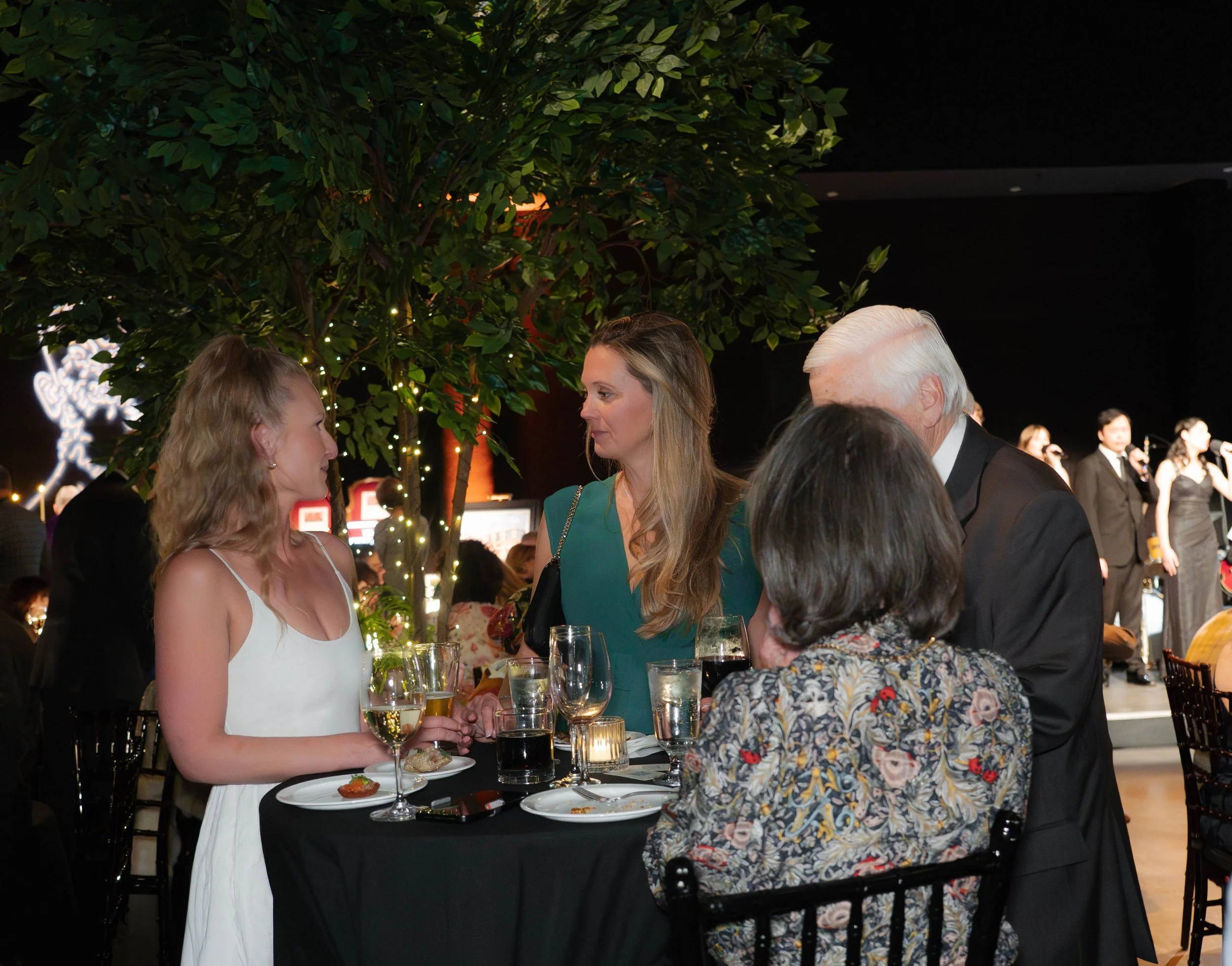 Four people gathered around an elegant black banquet table at a formal event, with a large indoor artificial tree decorated with small string lights in the background.