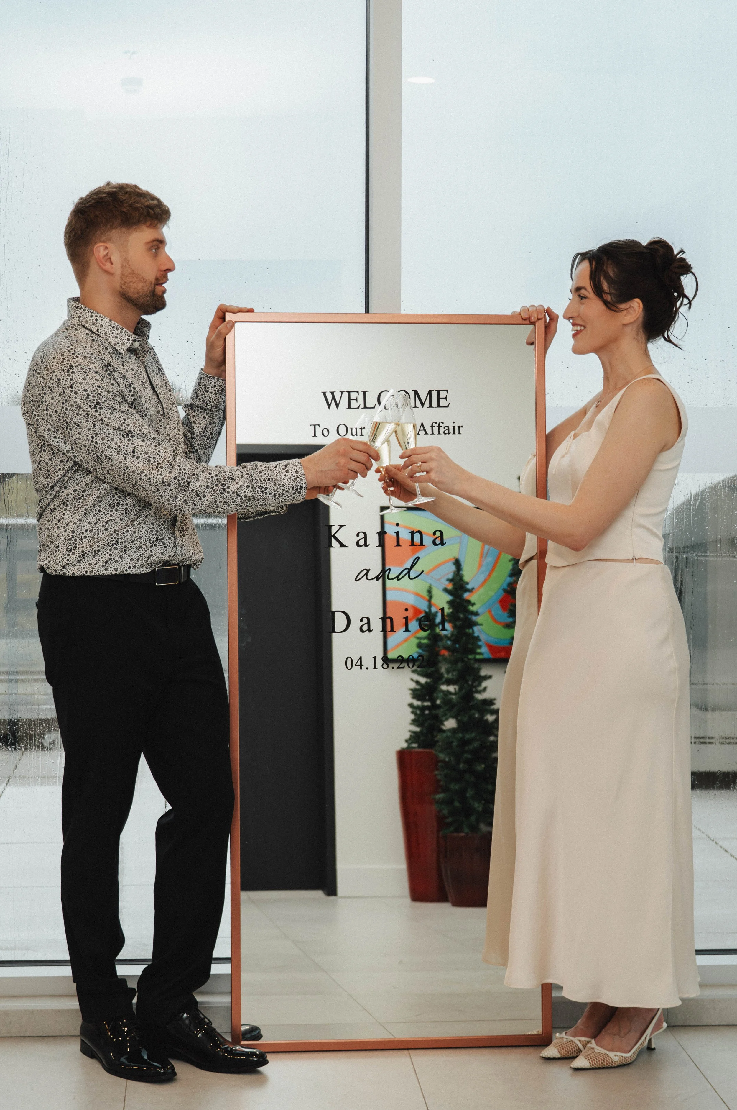 A man and woman are toasting with champagne glasses in front of a welcome sign that reads 'WELCOME To Our Wedding Affair Karina and Daniel 04.18.2023'. They are smiling and dressed formally, with a Christmas tree in the background.