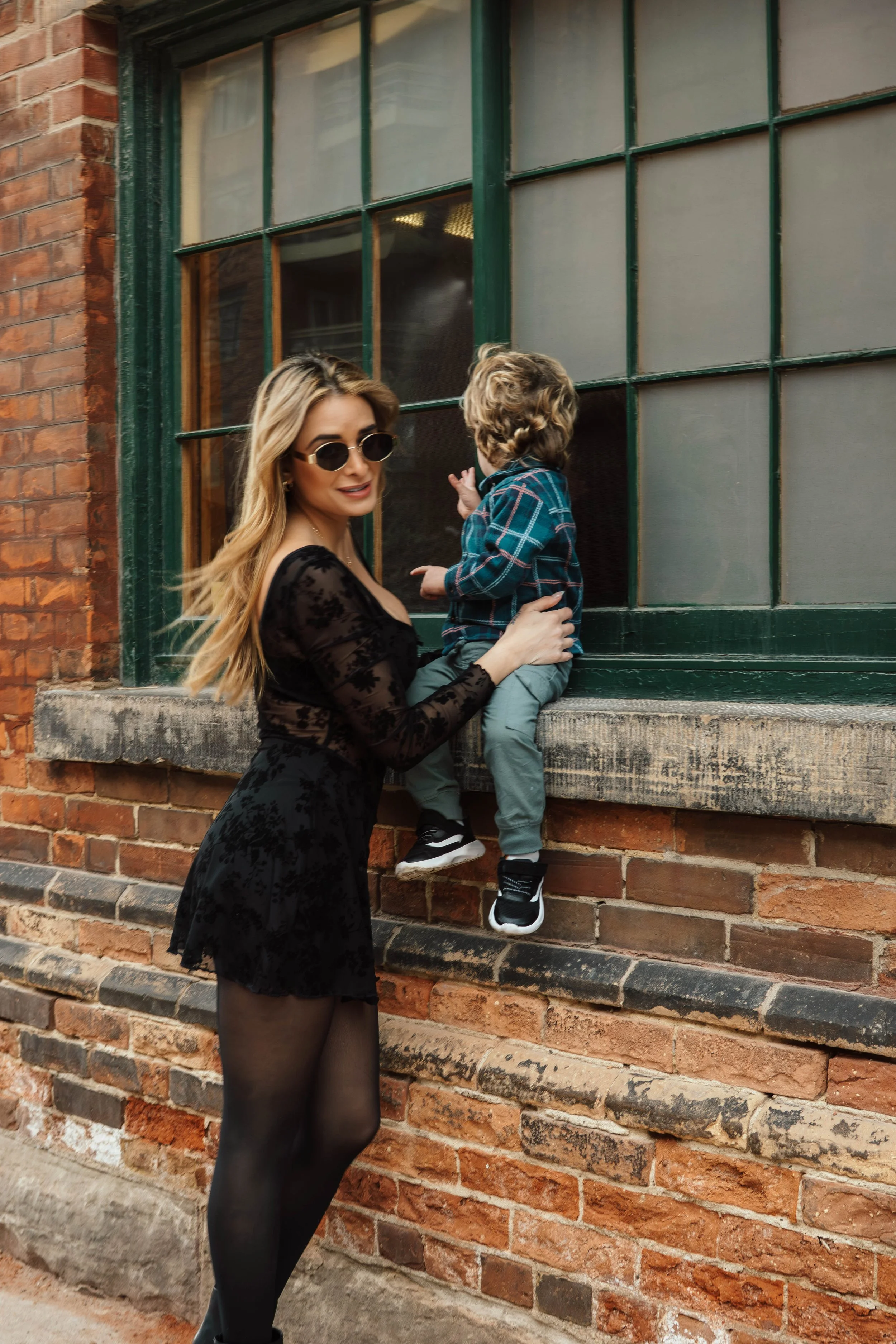 A woman with long blonde hair wearing sunglasses, a black lace dress, and black tights, standing next to a brick wall. She is holding a young boy with light brown curly hair, who is wearing a plaid jacket and gray pants, and is looking out a green-fr