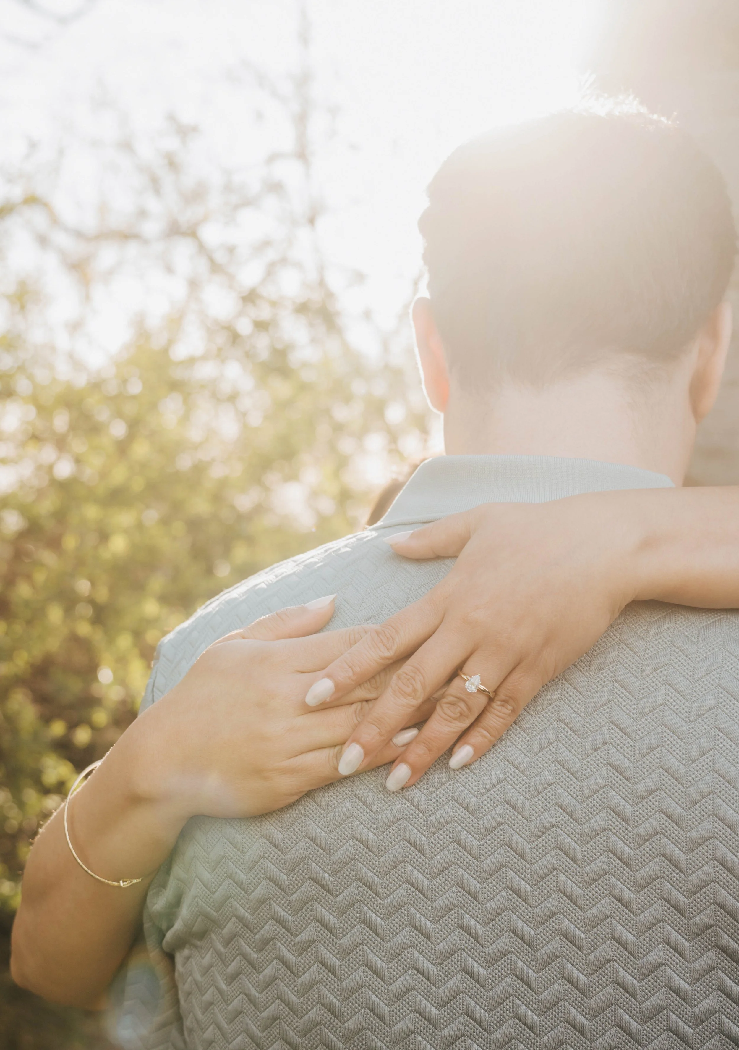 A woman with a diamond engagement ring, a gold bracelet, and a thin gold bracelet, hugging a man. The woman has a wedding ring and her nails are painted white. The photo is taken from behind and shows the back of their heads and arms, with a backgrou