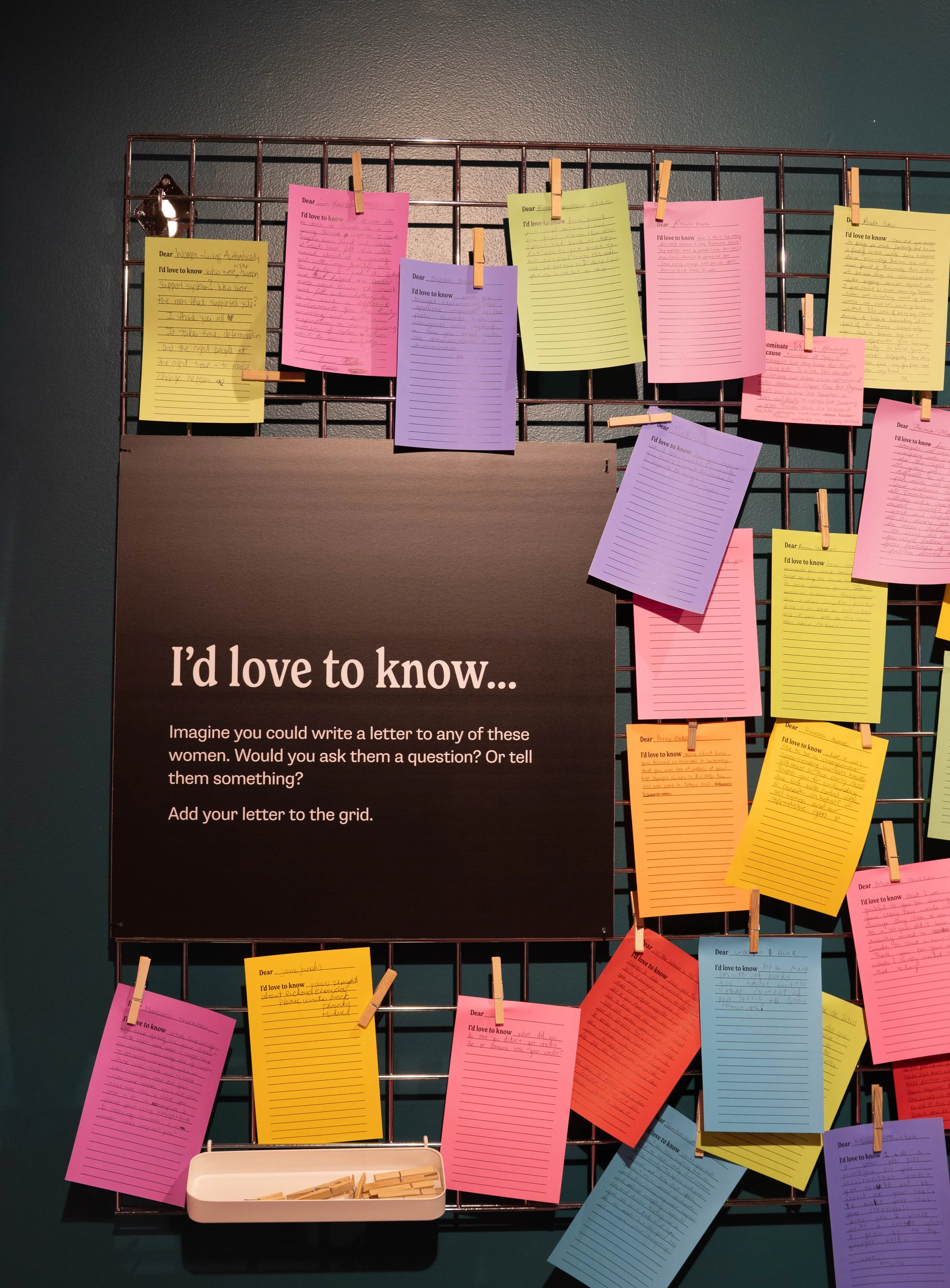 A display board with colorful lined paper notes clipped to a grid, titled 'I'd love to know...' with instructions to imagine writing a letter to women, asking questions or sharing thoughts, and adding the letter to the grid.