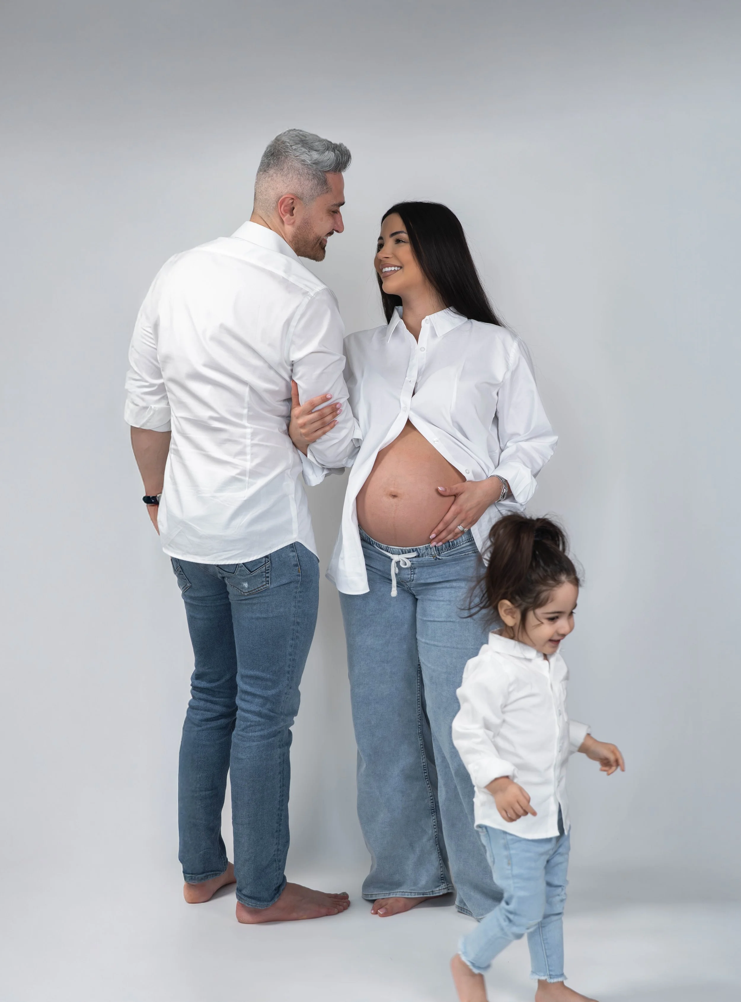 A happy pregnant woman with long black hair, wearing a white shirt and ripped jeans, holding her belly, standing next to a smiling man with short gray hair, wearing a white shirt and jeans, looking at the woman. Two young girls with dark hair are als