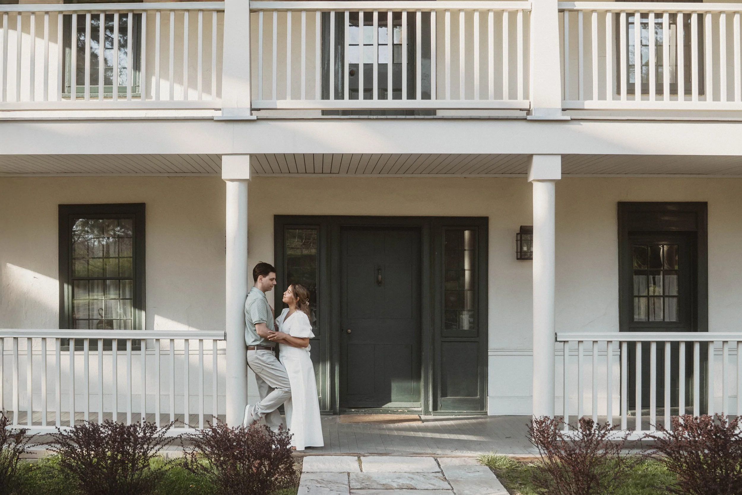 A couple standing on the porch of a white house, looking into each other's eyes, with the man leaning against a white pillar and the woman holding his hands.