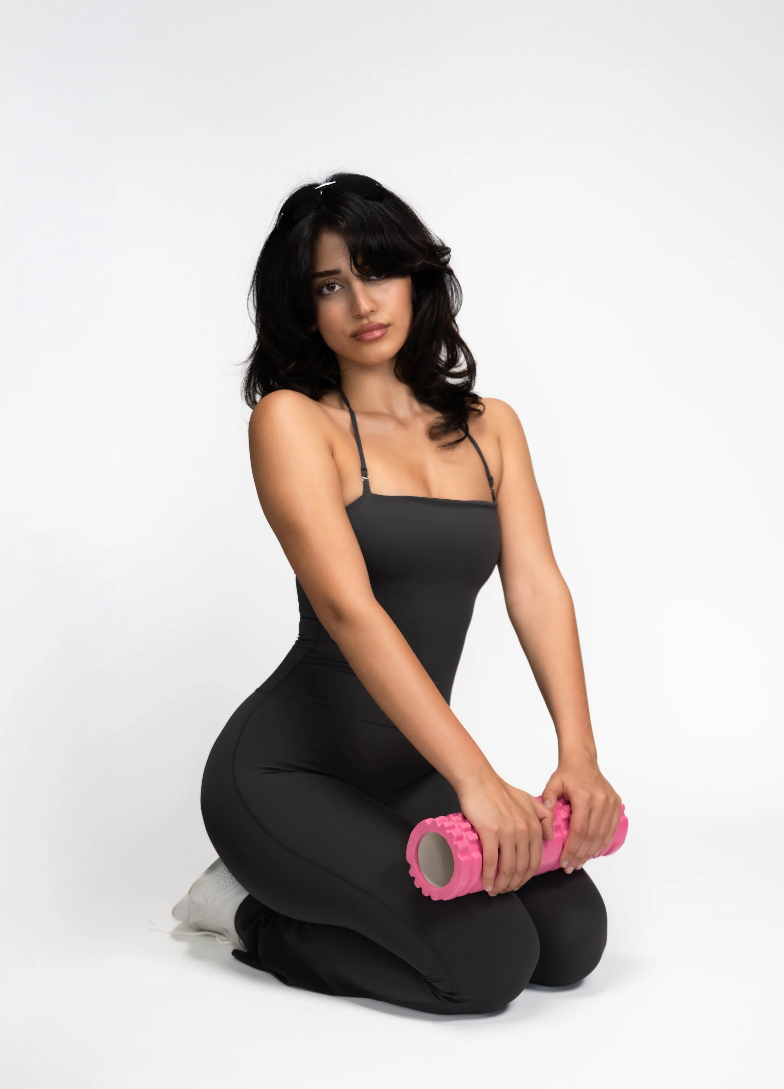 A woman kneeling on the floor, holding a pink foam roller with both hands, wearing a black tank top and black pants, with a white background.