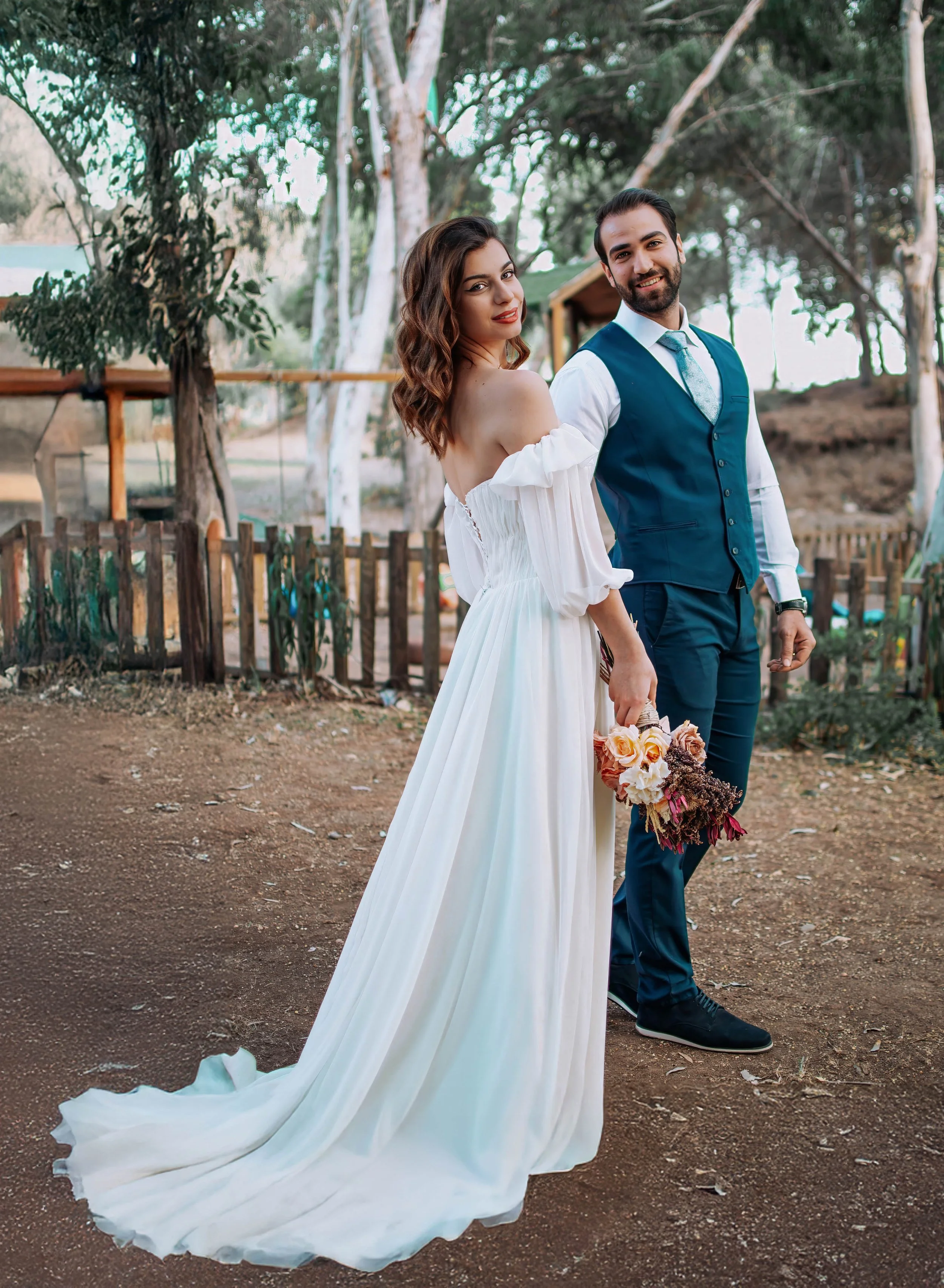 A bride and groom standing outdoors in a park-like setting with trees and a fence in the background. The bride is wearing a white off-shoulder wedding dress and holding a small bouquet, while the groom is dressed in a blue vest and suit with a white 