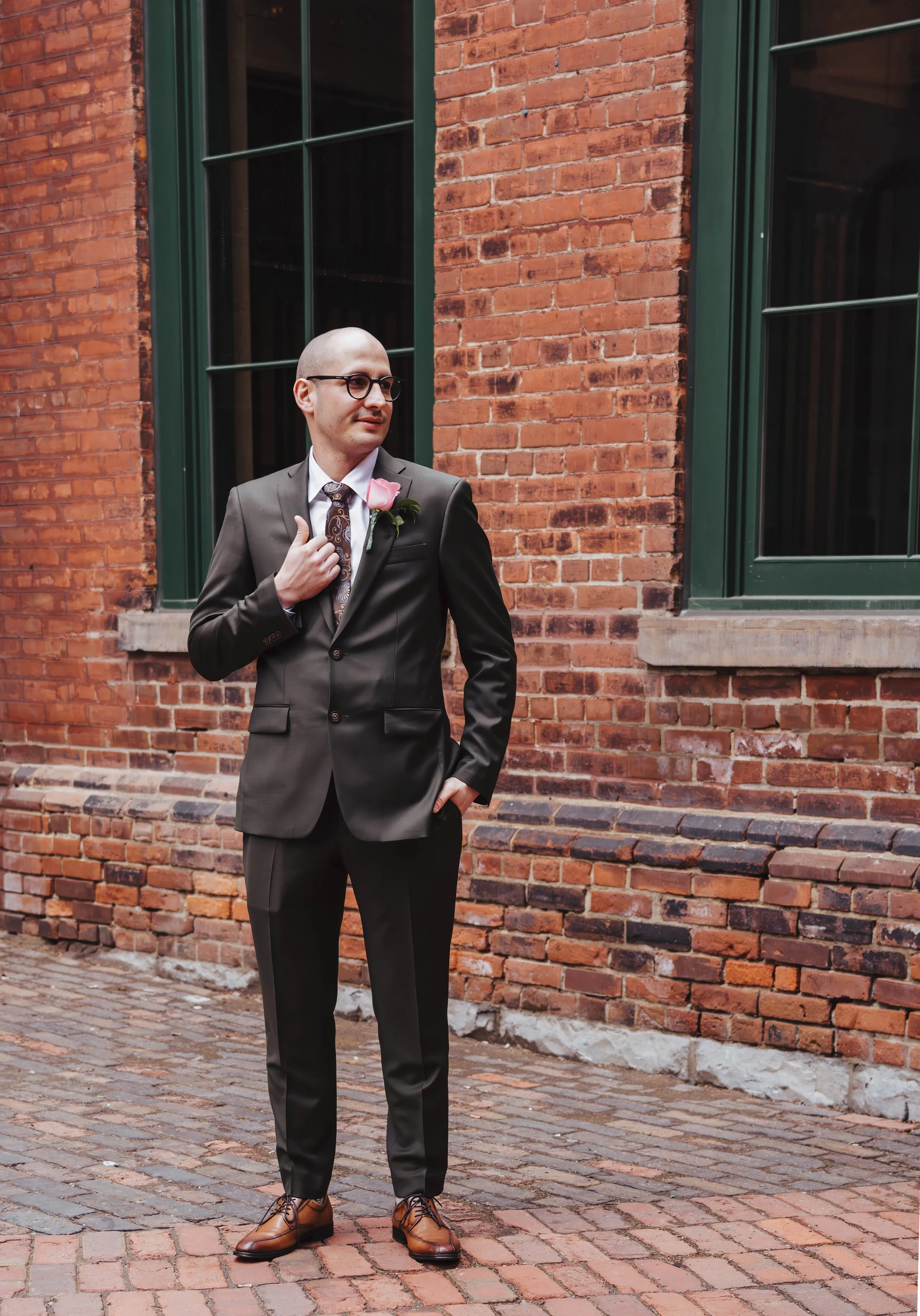Man in a dark suit and glasses standing outside on a brick street, adjusting his suit lapel, with a pink flower boutonniere, in front of a red brick building with large green window frames.