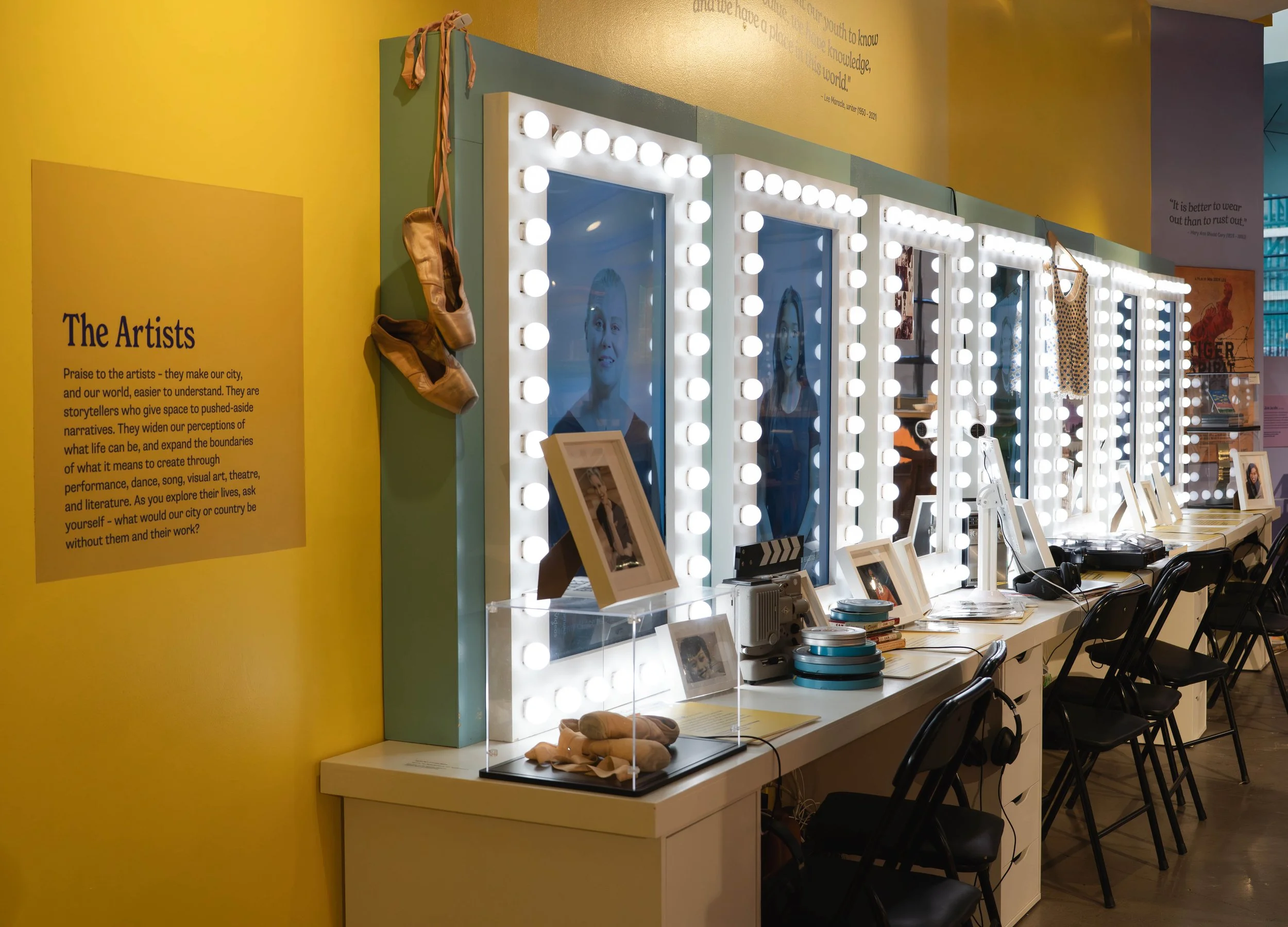 A row of illuminated mirrors with light bulbs, desk, and chairs in front of a yellow wall with informational text about artists.