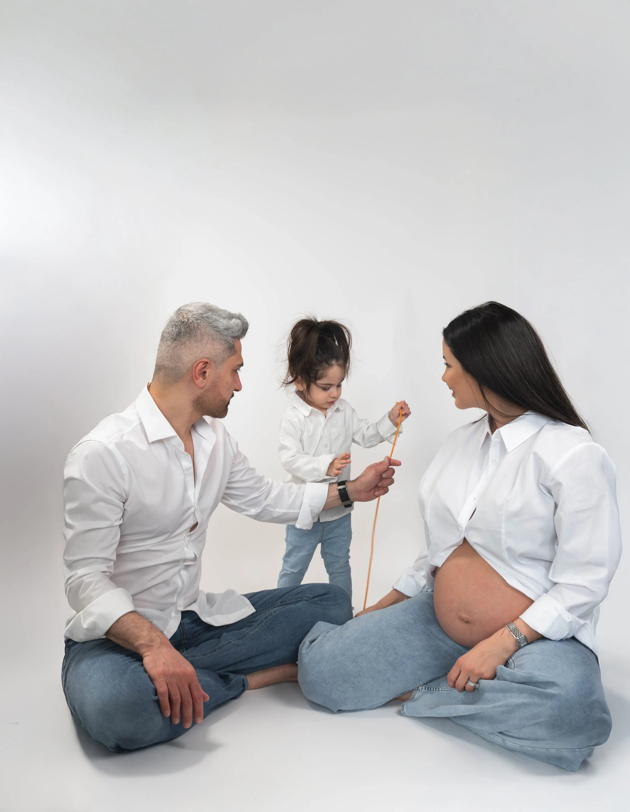 A pregnant woman sitting on the floor with her partner, a man, and their young daughter. The woman is pregnant and smiling, the man is holding the daughter, who is standing on her lap and holding a toy. The family is dressed in white shirts and blue 