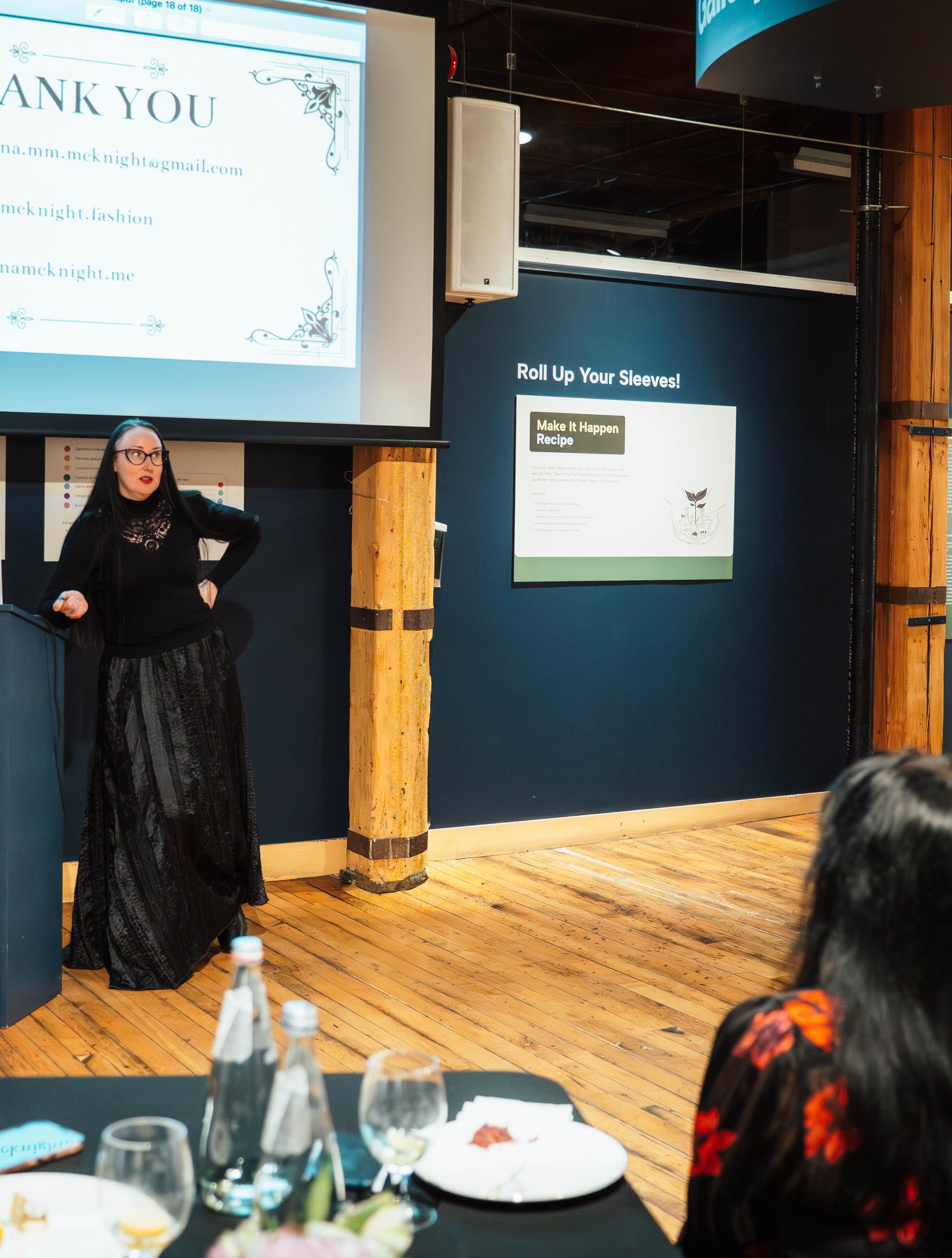 A woman giving a presentation in a conference room, with a large screen displaying a 'Thank You' slide and smaller screens with informational slides, wooden accents, and a table with bottles and dishes in the foreground.
