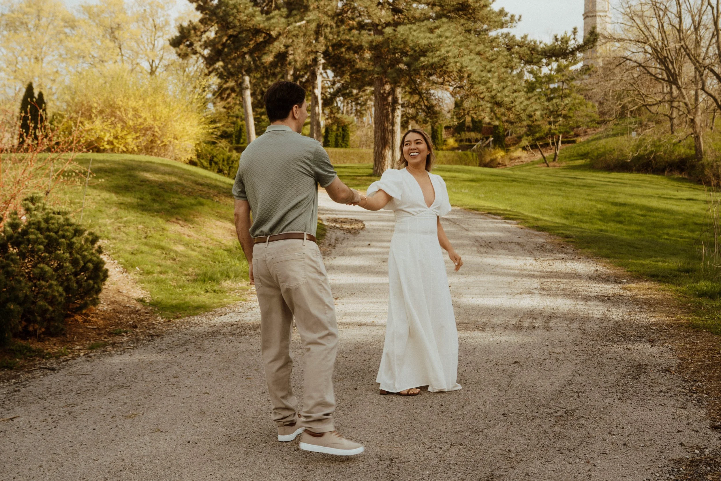 A smiling woman in a white dress holding hands with a man in casual clothing, standing outdoors on a path amidst trees and greenery during the daytime.