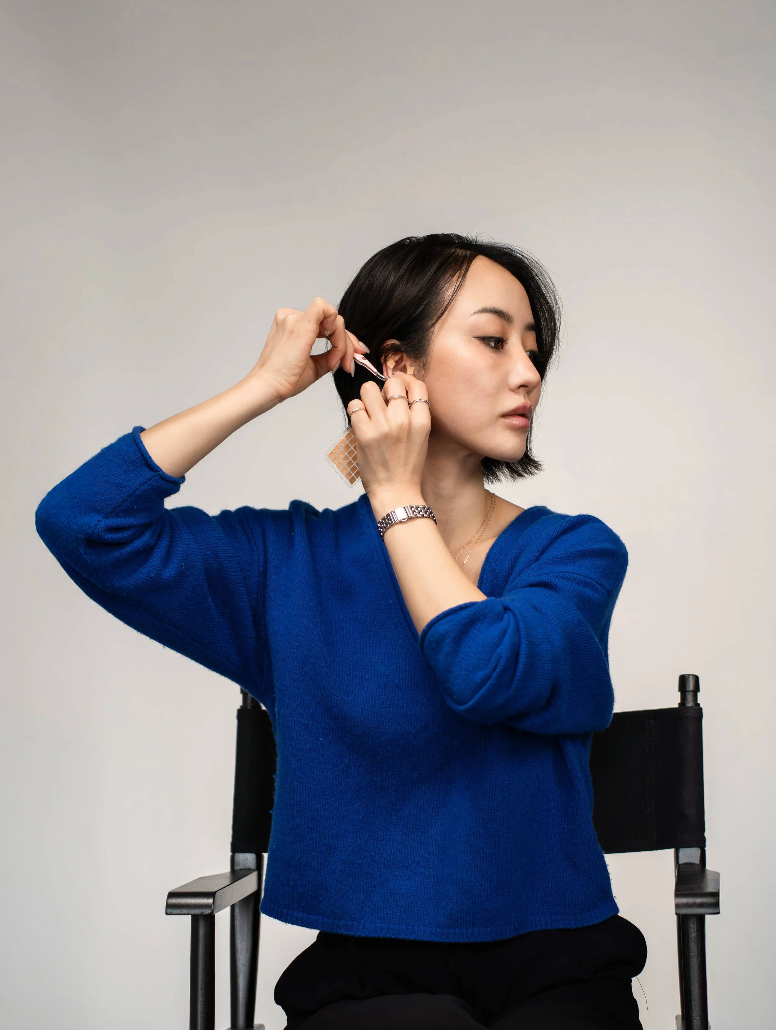 A woman with black hair wearing a blue sweater is sitting on a chair and attaching a hair extension to her ear.