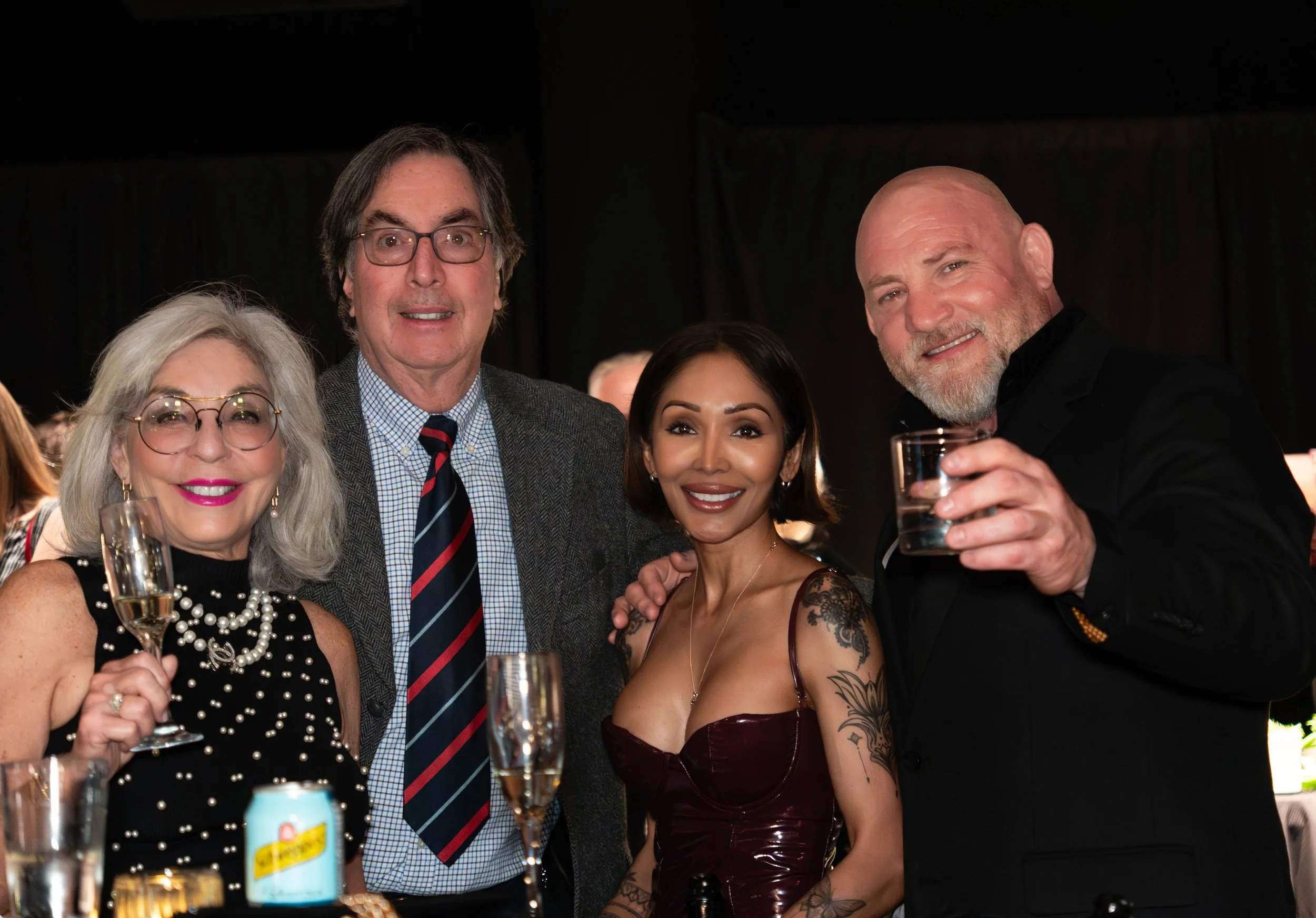 Four people smiling at a party, holding drinks, dressed in formal attire, with two women and two men.