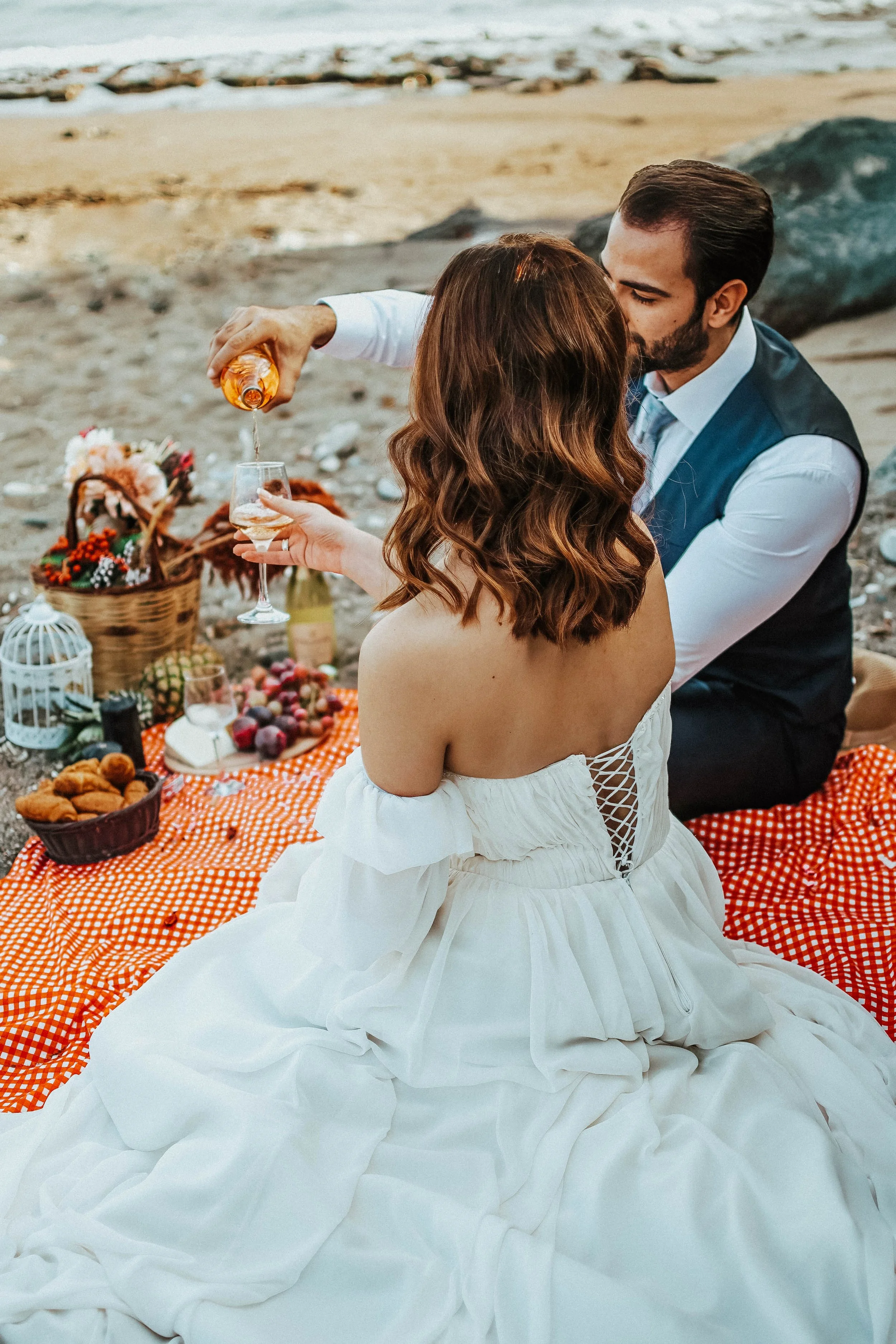 A couple having a romantic picnic on the beach, with the man pouring a drink into a woman's glass, surrounded by a picnic blanket and food.