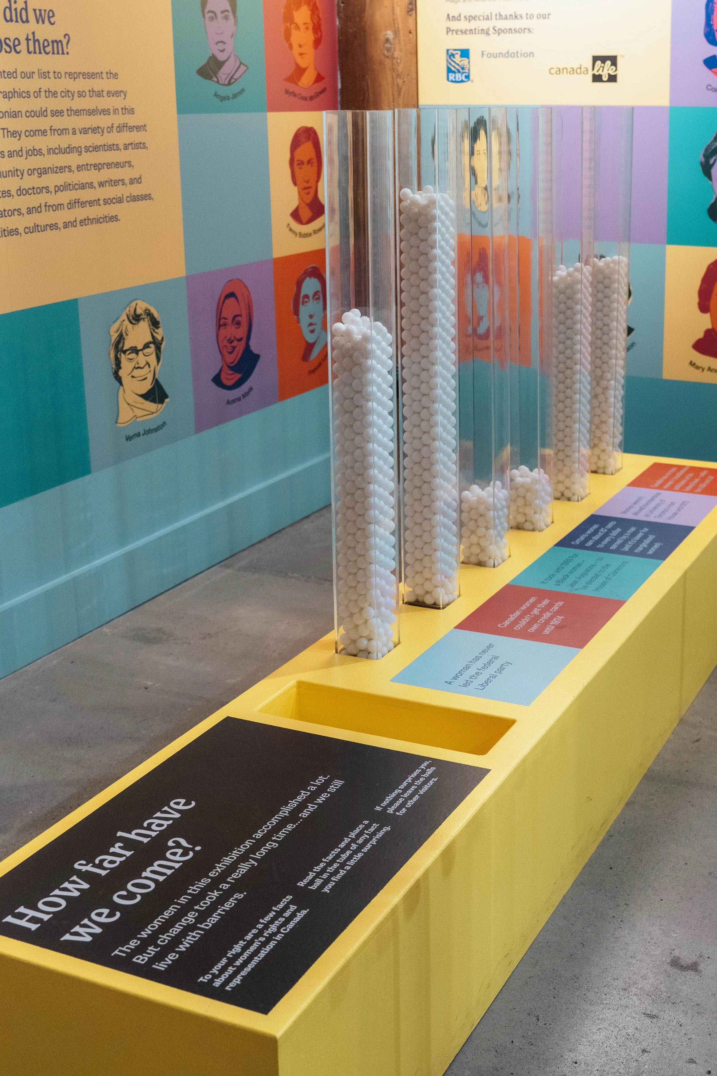 An interactive exhibit with five transparent tubes filled with white balls, set on a yellow display table. Behind, there are colorful portraits of women and informational panels about women's achievements and history.