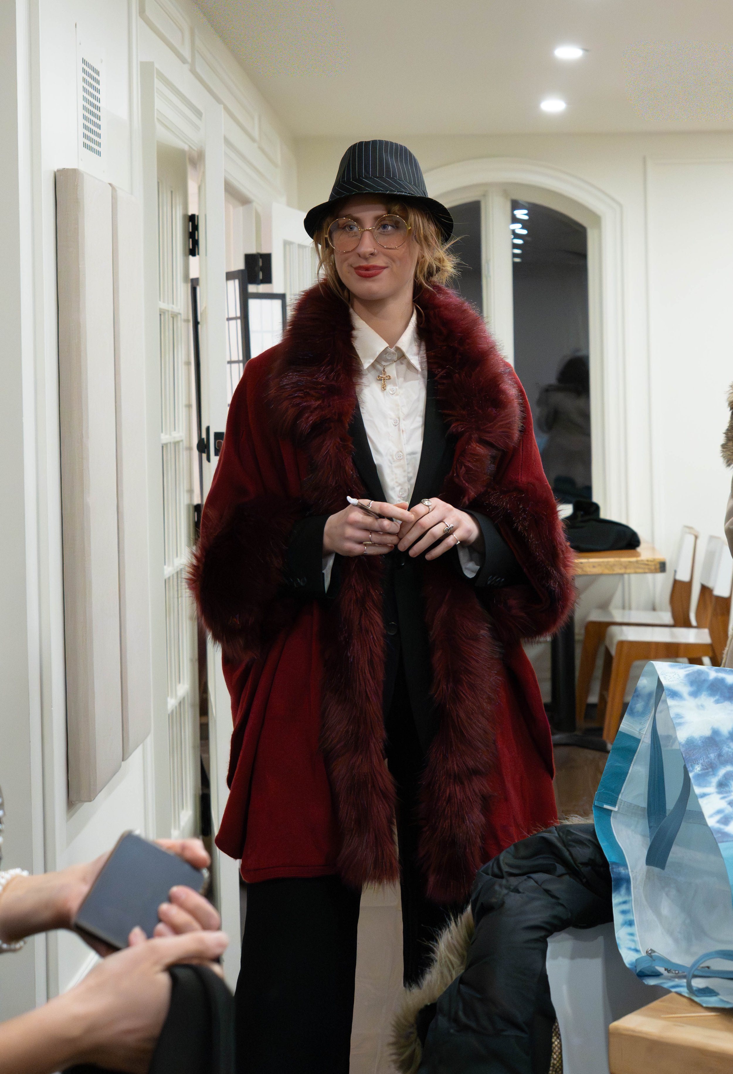 A woman wearing a striped hat, glasses, and a red fur coat with a black lapel stands indoors, holding a pen and smiling. There are people and objects around her, including a person holding a phone, a bag on the table, and chairs.
