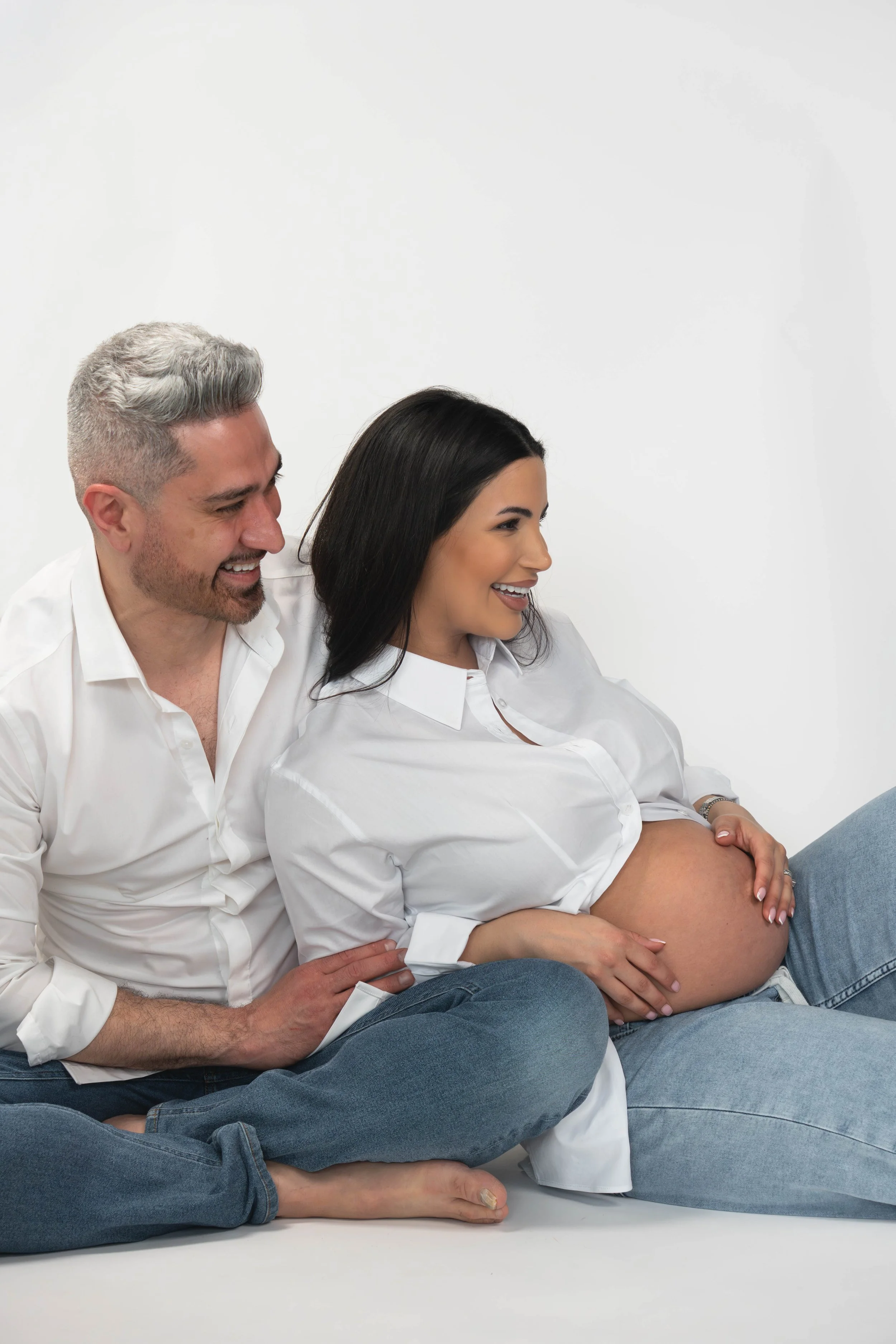 A pregnant woman with long dark hair sitting on the floor, smiling, with her hands on her belly. She is wearing a white shirt and blue jeans. A man with short, gray hair and a beard, also smiling, is sitting behind her with his arms around her, weari