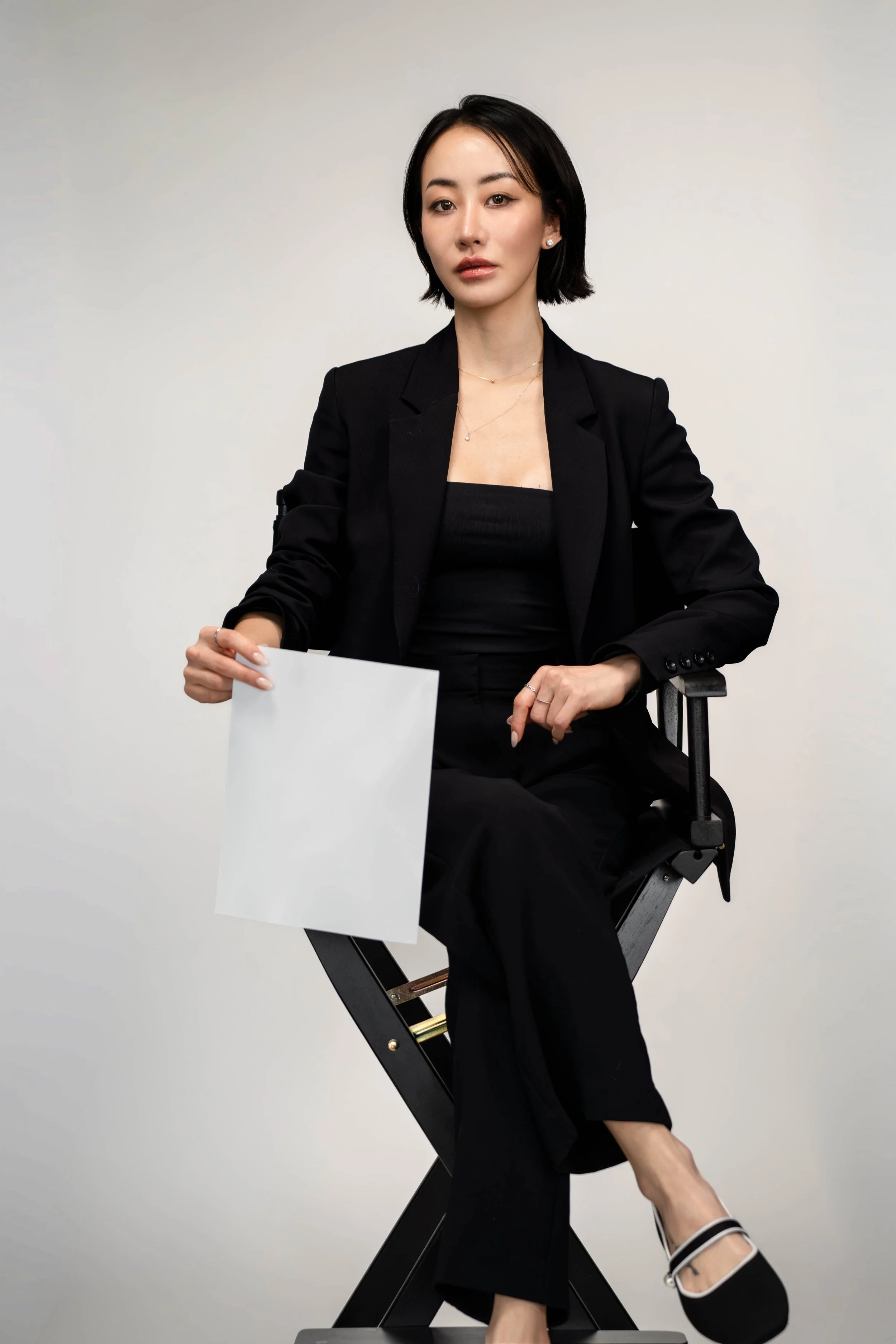 A woman with dark hair wearing a black blazer and pants, sitting on a chair, holding a blank white paper, against a plain light-colored background.