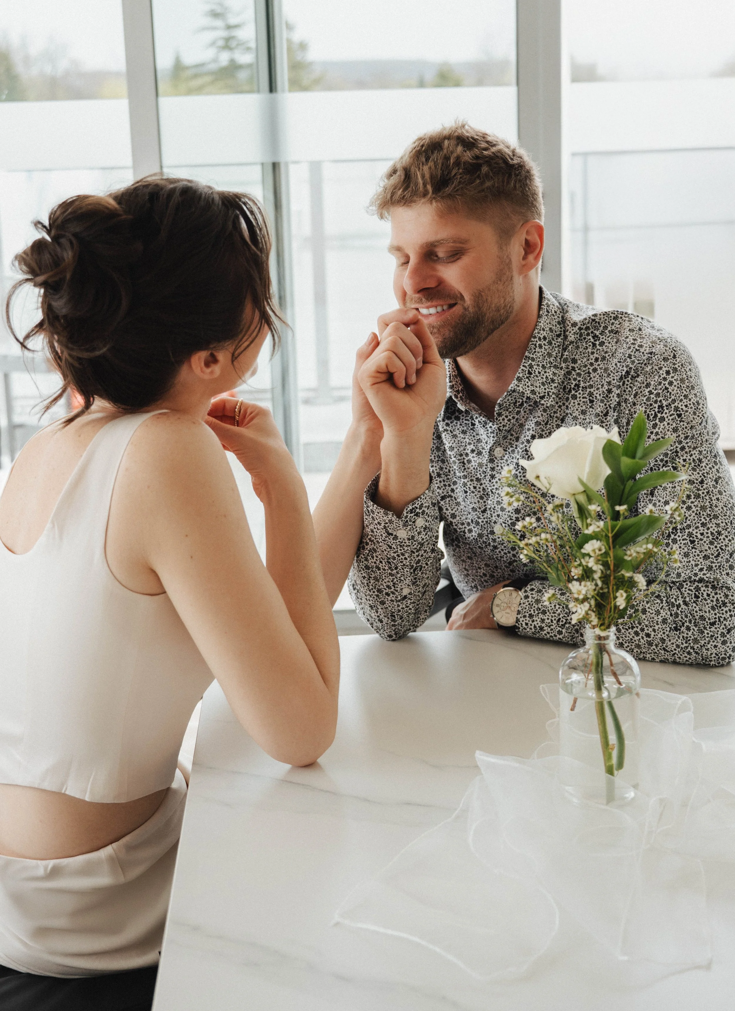 A man and woman are seated at a white table, with the man offering the woman a flower. The woman is smiling and looking at the man. The table has a vase with white flowers and green leaves, wrapped in tissue paper. The background shows large windows 