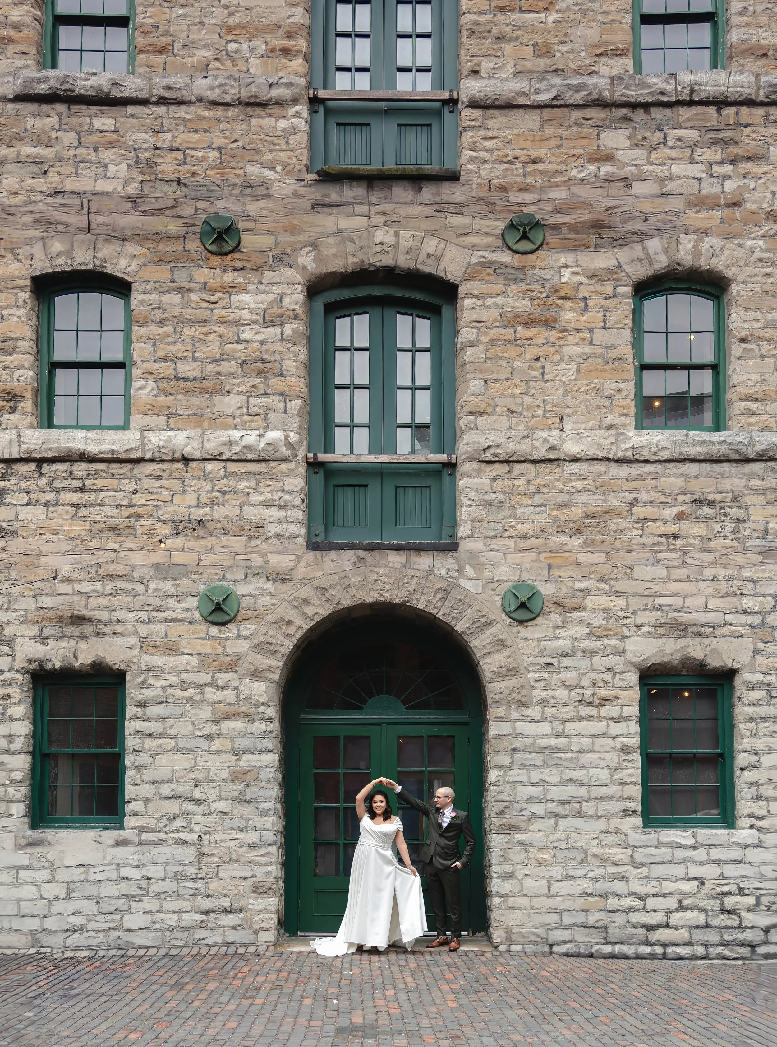 A bride and groom pose for a wedding photo in front of a rustic stone building with green doors and windows, with the groom twirling the bride.