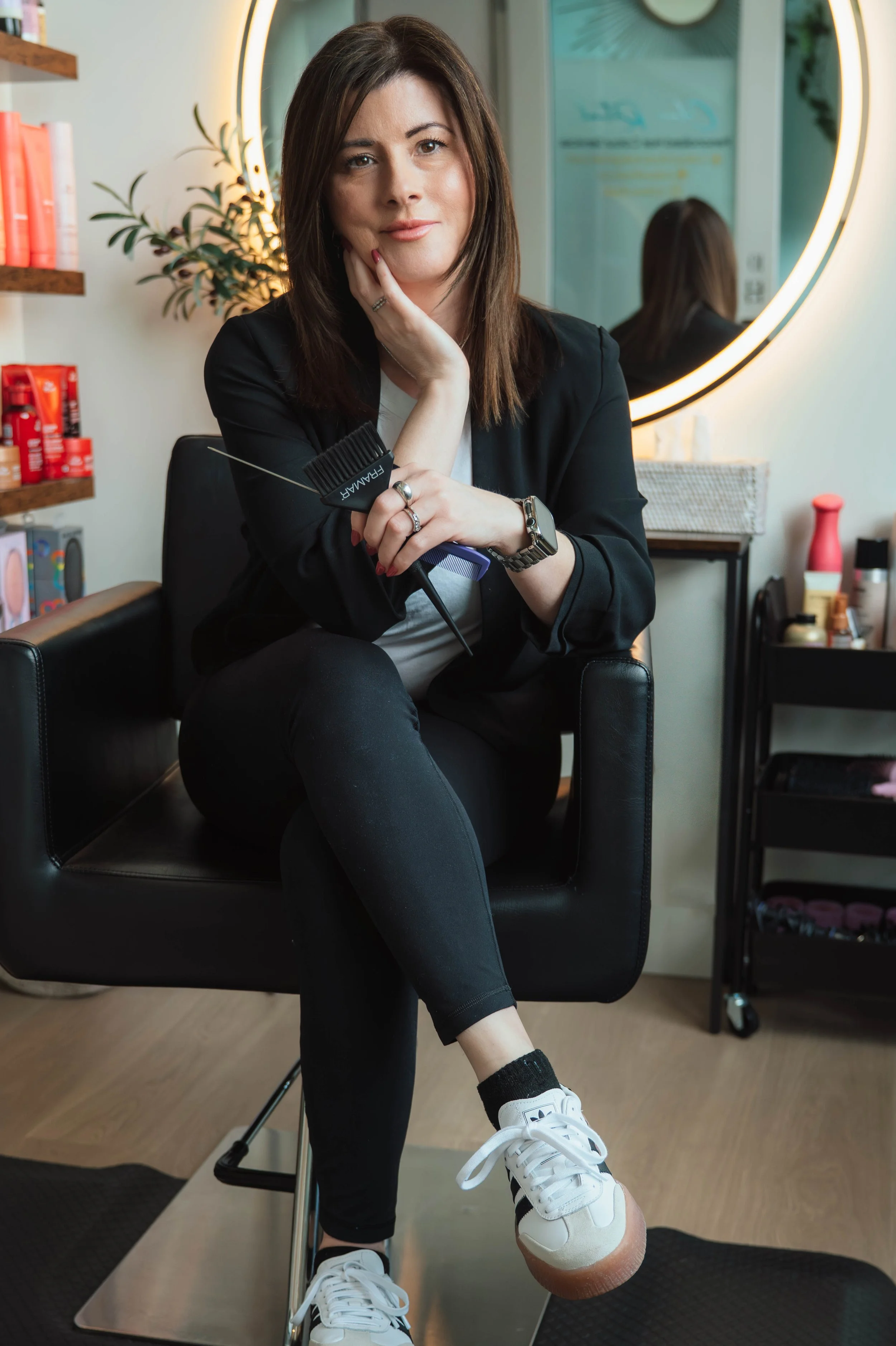 A woman with brown hair sitting in a salon chair holding a hairbrush and a comb, wearing a black jacket, white shirt, black pants, white sneakers, and an Apple Watch, with a salon mirror and shelves of hair products in the background.