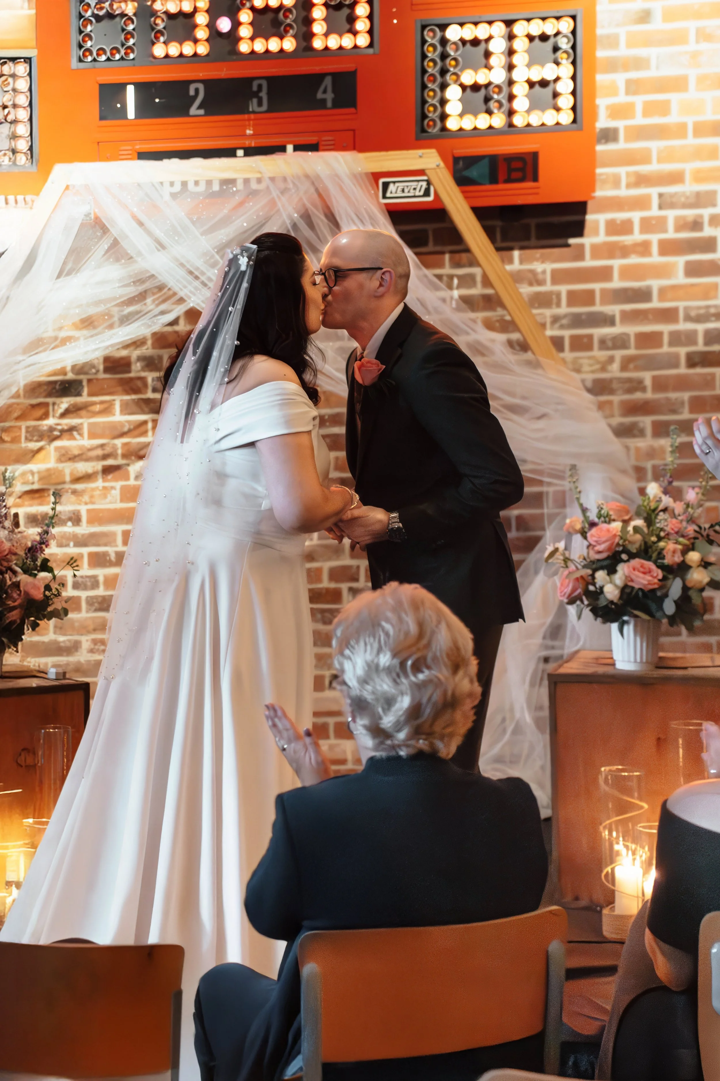 Couple getting married, kissing during their wedding ceremony, with guests clapping, decorated with floral arrangements and a brick wall backdrop.