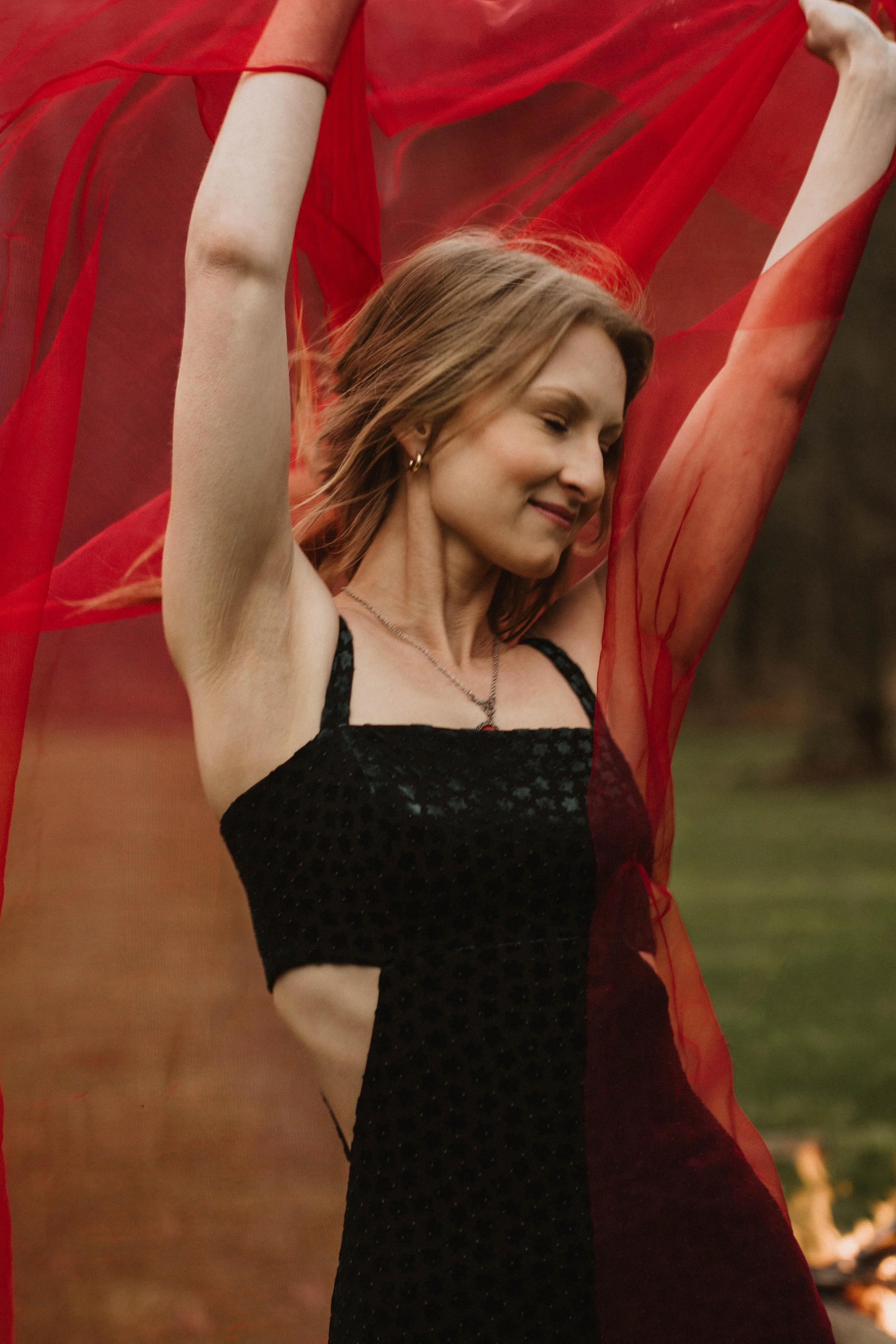 A woman with short light brown hair, wearing a black dress with cutouts, holding a flowing red fabric above her head outdoors.