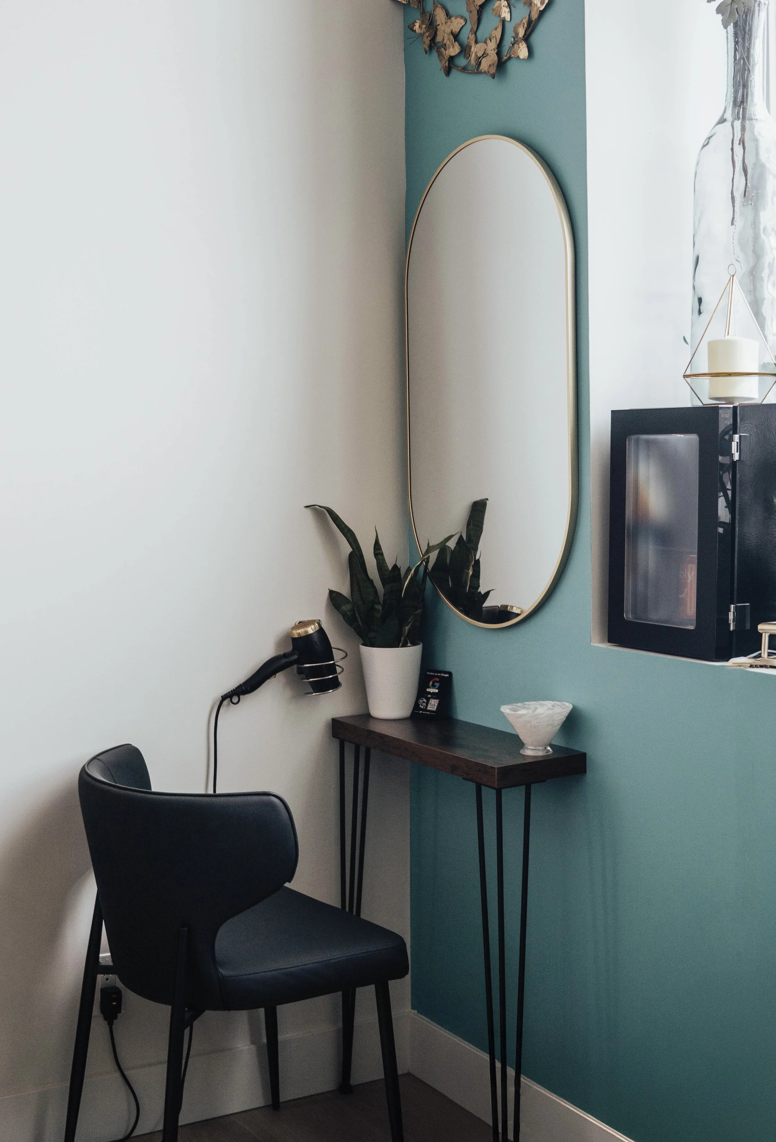 A small workspace corner with a black chair, a wooden desk with metal legs, a potted plant, a mirror on a teal accent wall, and a window with a black microwave and decorative candle holder.