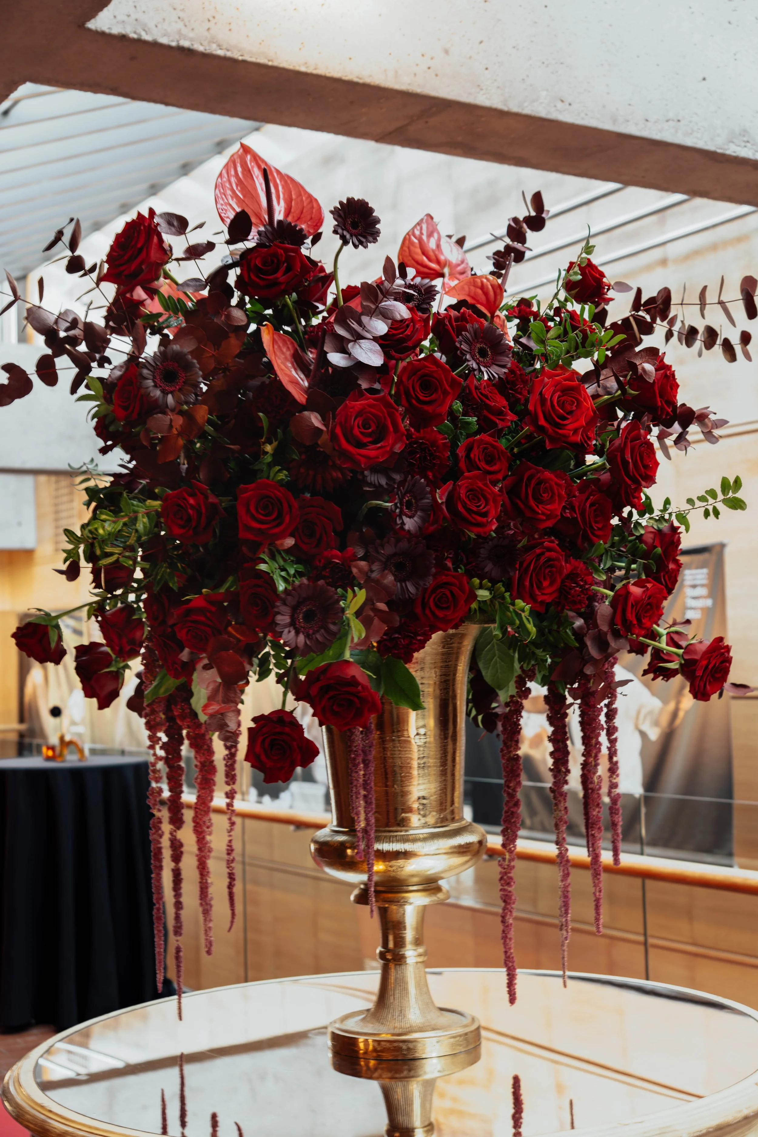 A large bouquet of red roses, dark anemones, and dark foliage in a shiny gold vase placed on a glass table.