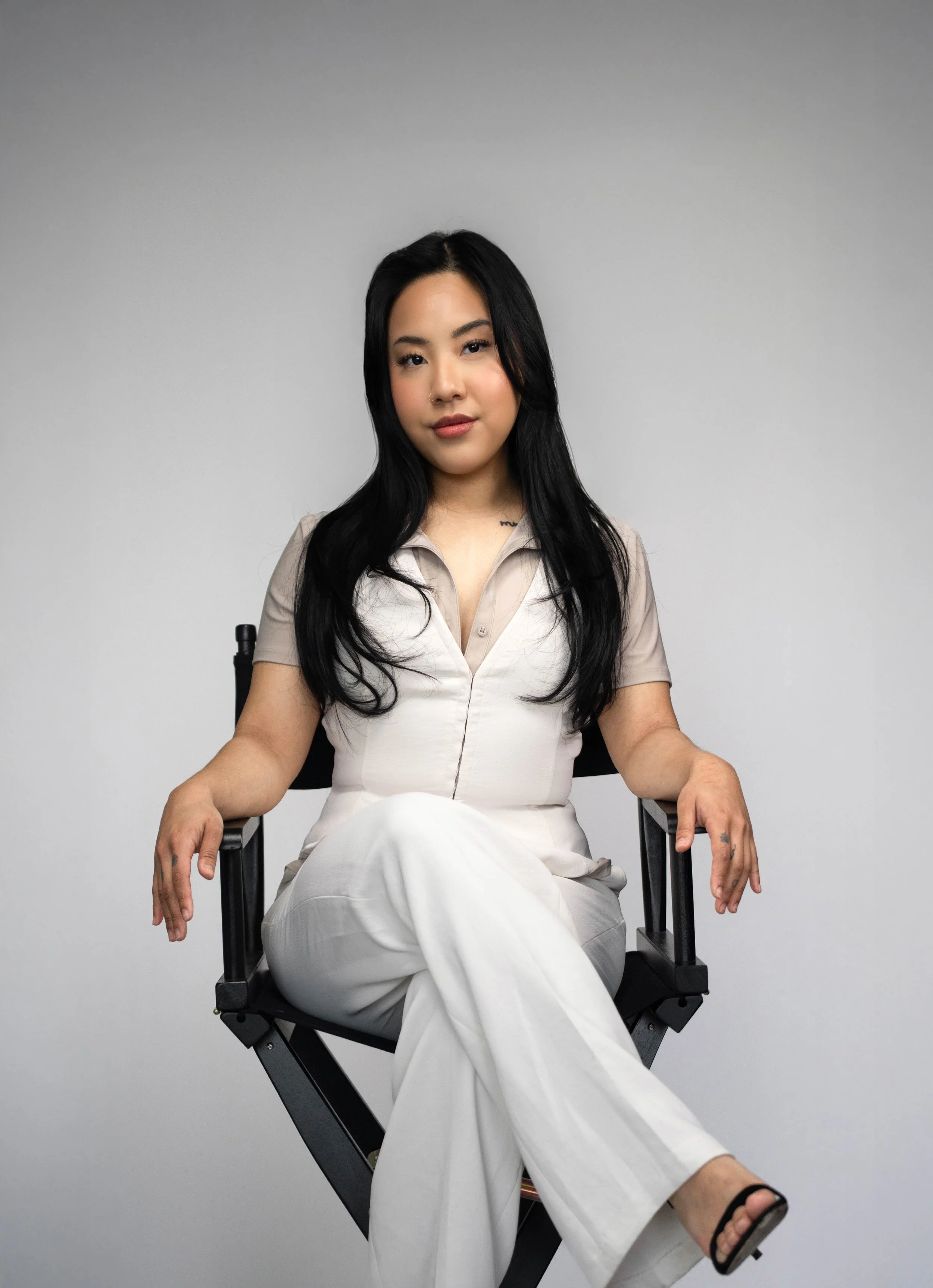 A woman with long black hair sitting on a director's chair against a plain gray background, wearing a beige short-sleeve shirt, white pants, and black sandals.