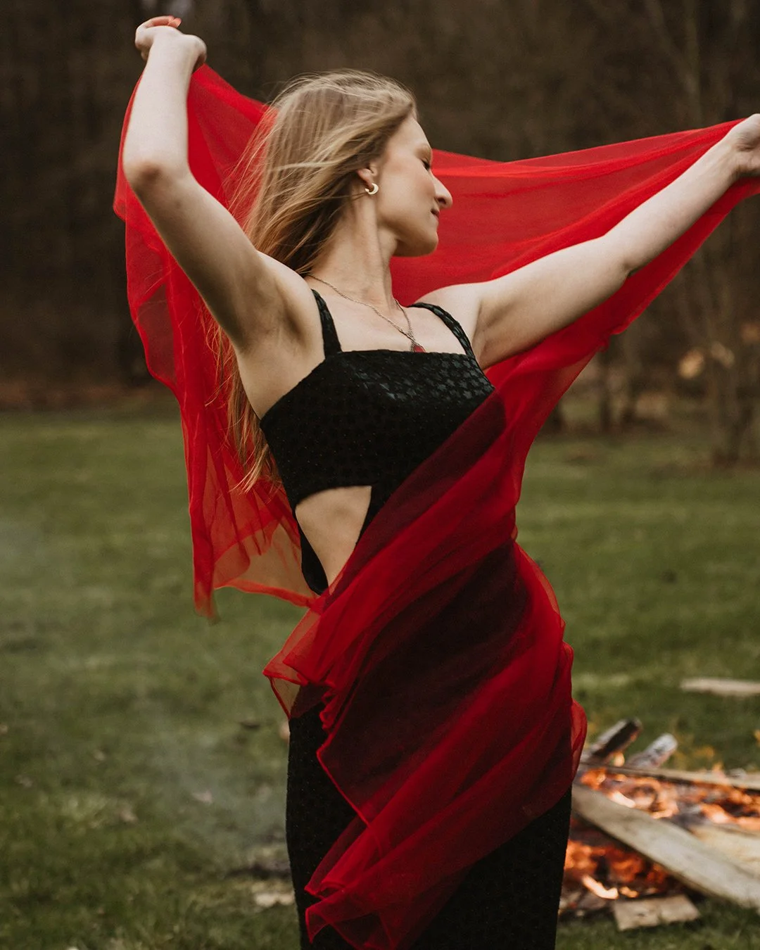 A woman in a black dress and red sheer shawl is standing outdoors with arms raised, holding the shawl open behind her, with a blurred background of trees and grass.