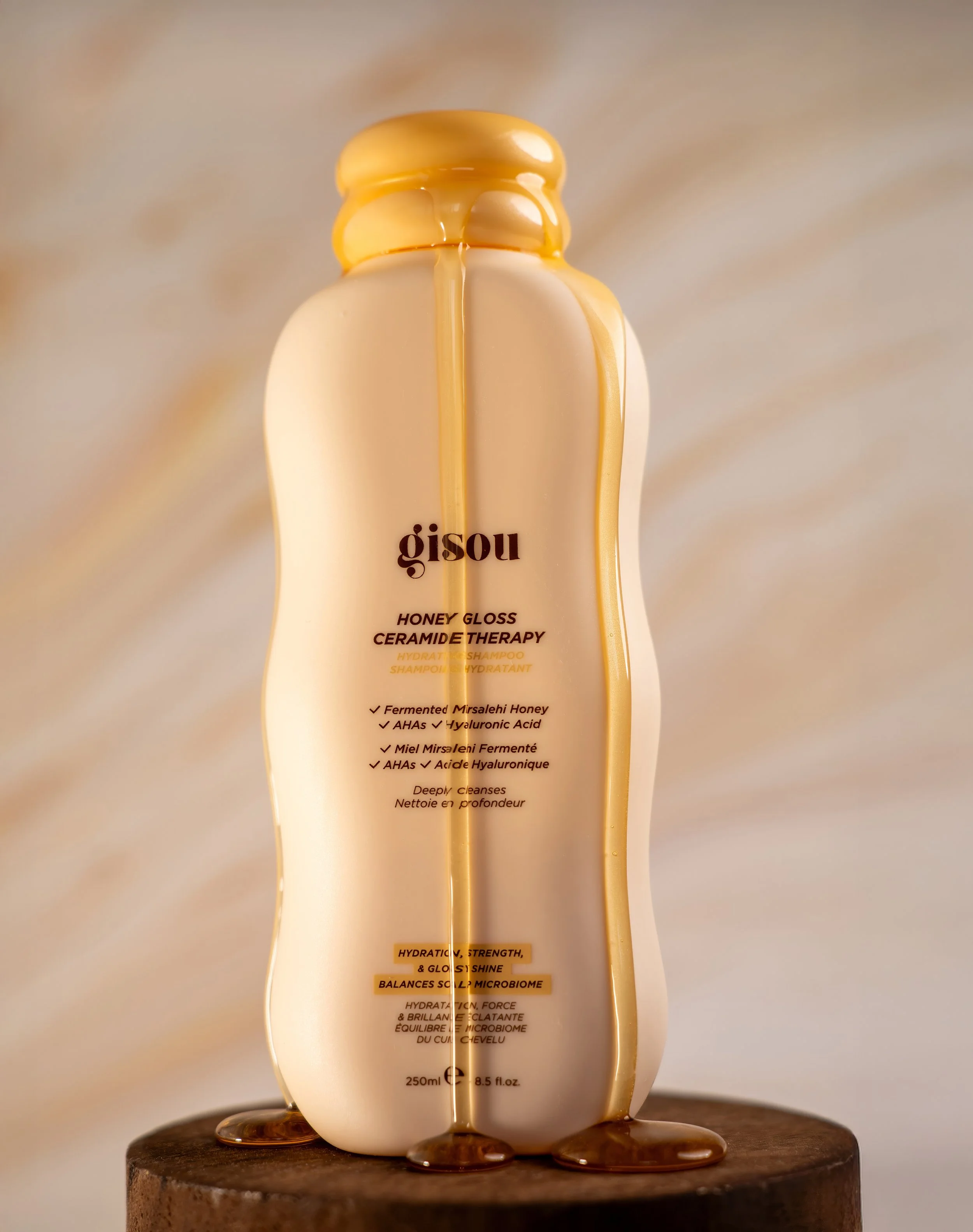 Bottle of Gisou Honey Gloss Ceramide Therapy shampoo with honey and hyaluronic acid, standing on a wooden surface with a blurred background.