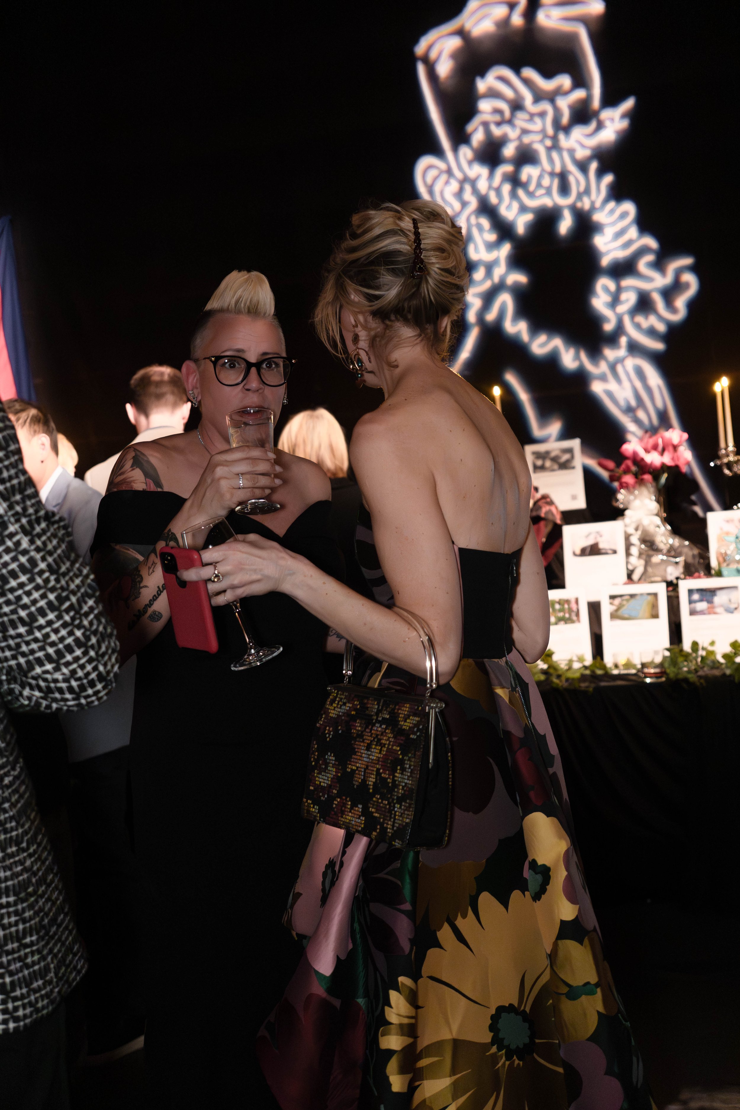 People at an indoor event, including two women in conversation, one holding a drink, with a decorated table and illuminated artwork in the background.