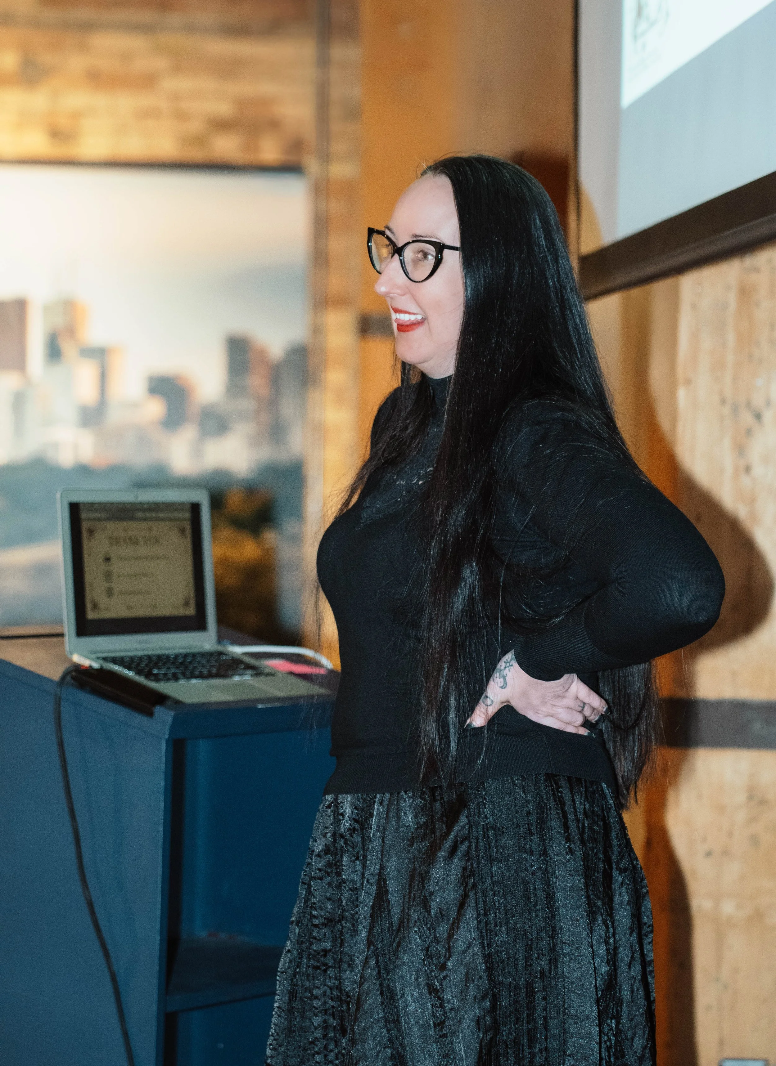 A woman with long black hair, glasses, and tattoos, standing with her hands on her hips in front of a presentation screen, during a presentation or lecture.