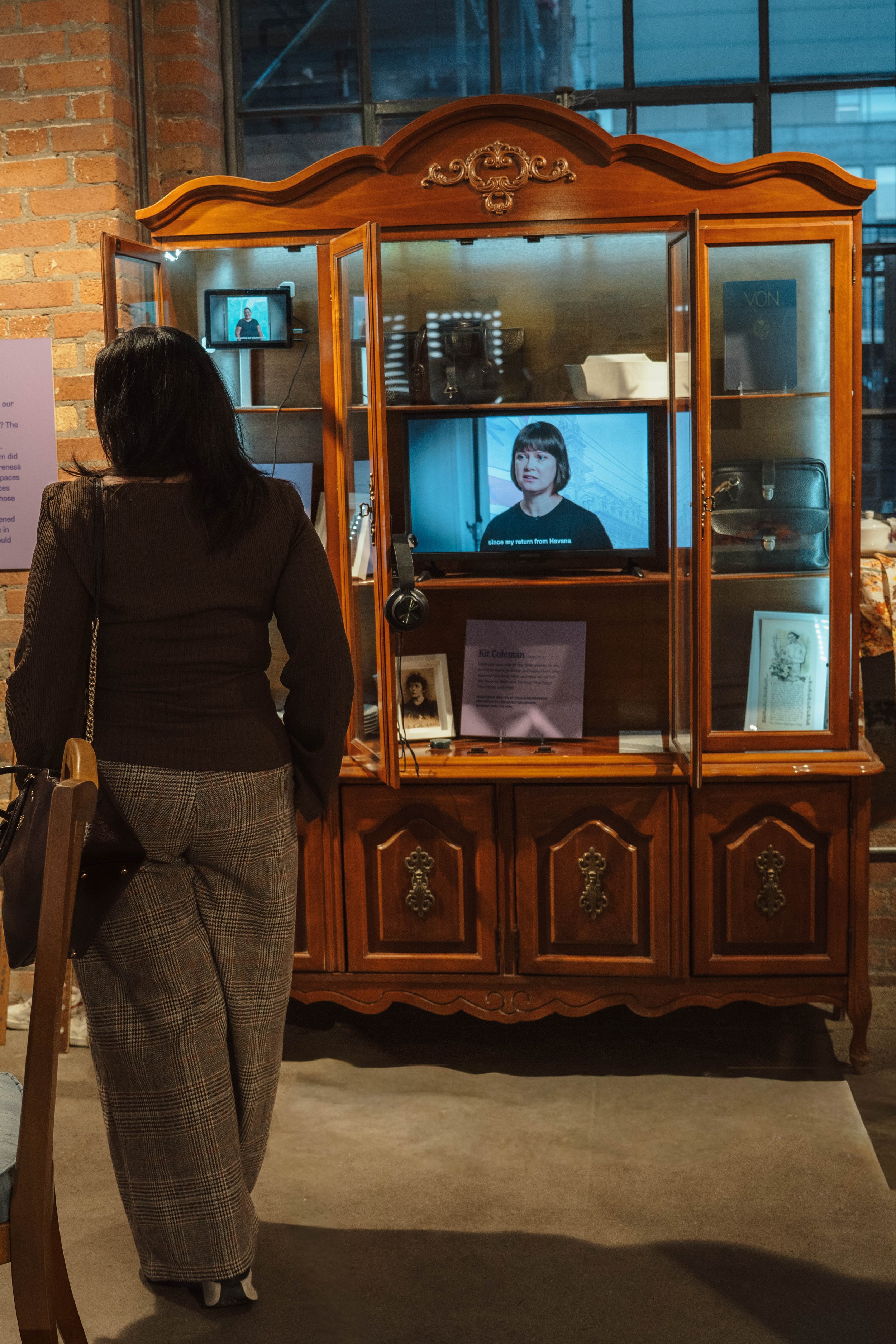 A woman stands in front of an antique wooden display cabinet with glass doors, watching a television screen inside the cabinet. The cabinet contains framed photos, a sign, and bags, with a brick wall and large window behind it.