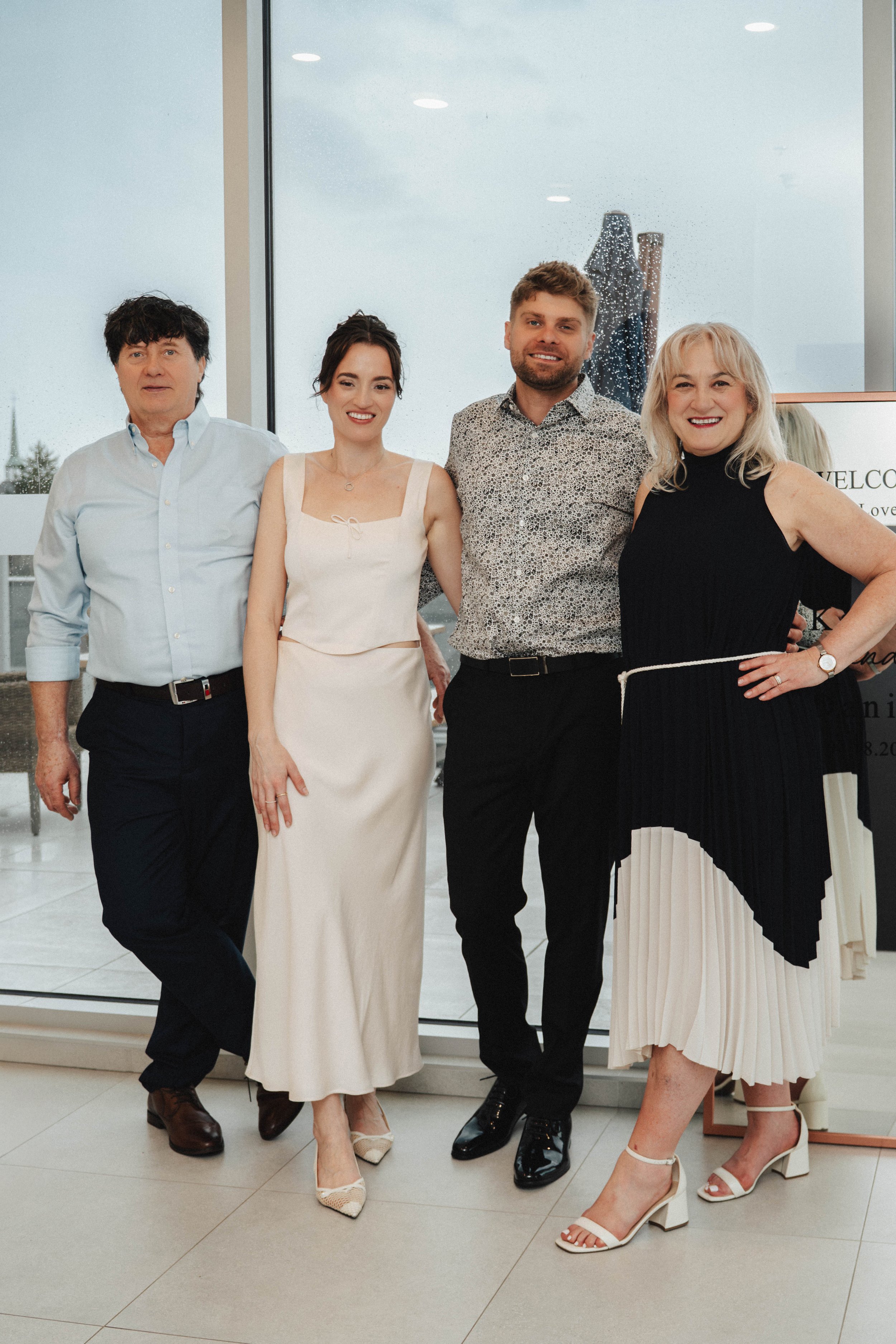 Group of four people posing indoors near a glass wall, with three women and one man dressed in formal attire, smiling and standing close together.