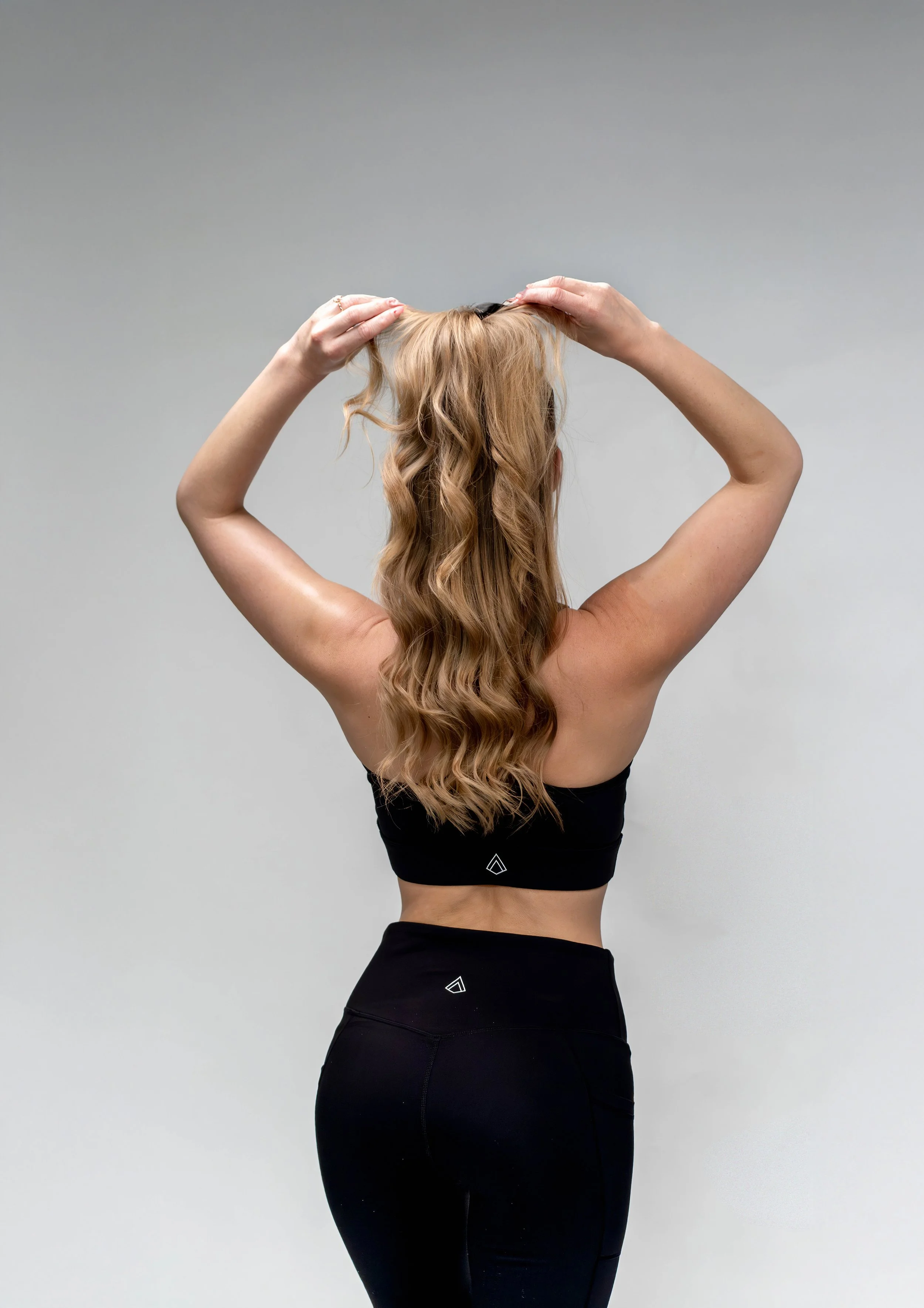 A woman in a black sports bra and leggings stands with her back to the camera, holding her long, wavy blonde hair up with both hands except her face.
