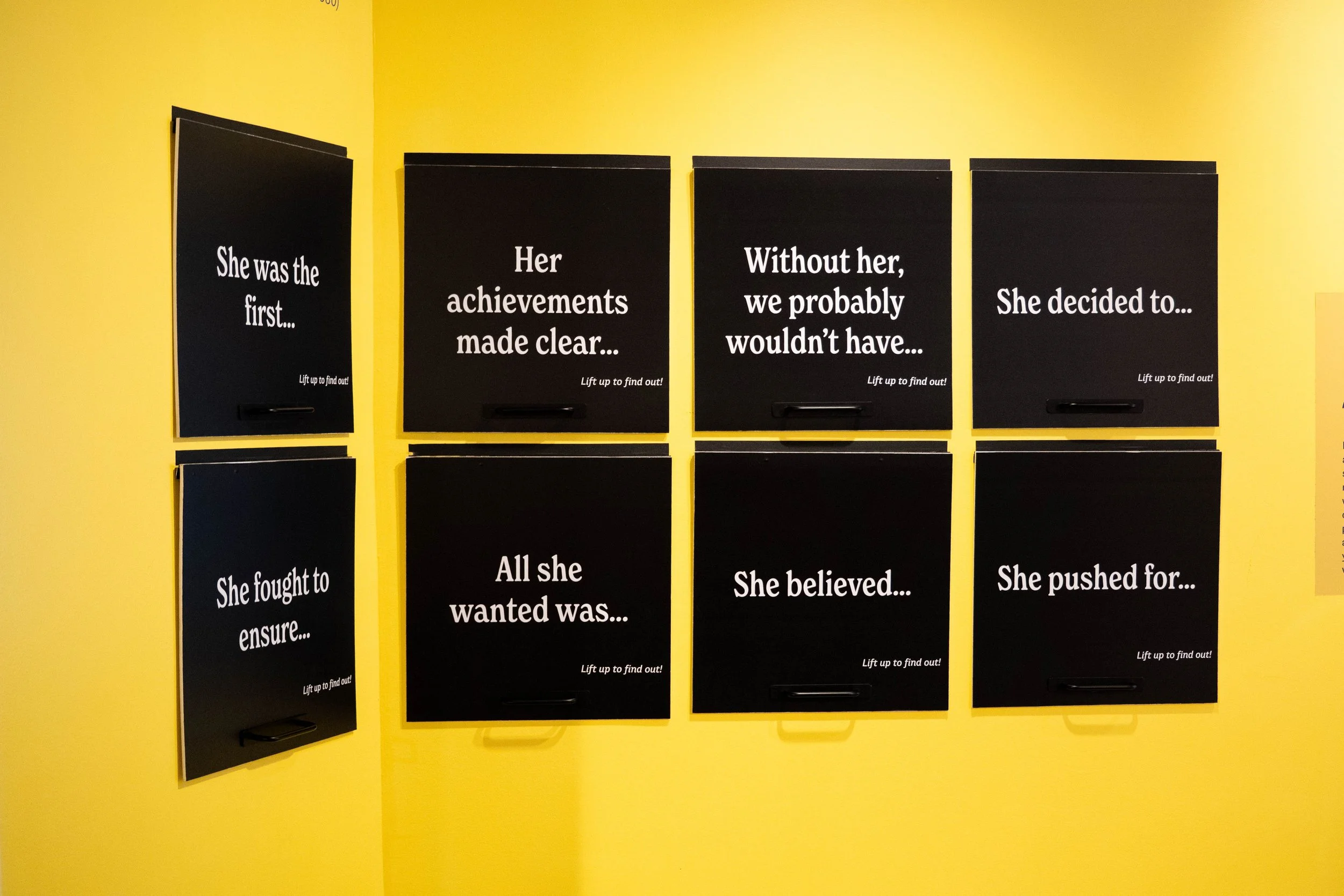 Eight black message boards with white text are mounted on a yellow wall in a 2x4 grid. The messages read: 'She was the first...', 'Her achievements made clear...', 'Without her, we probably wouldn’t have...', 'She decided to...', 'She fought to ensur