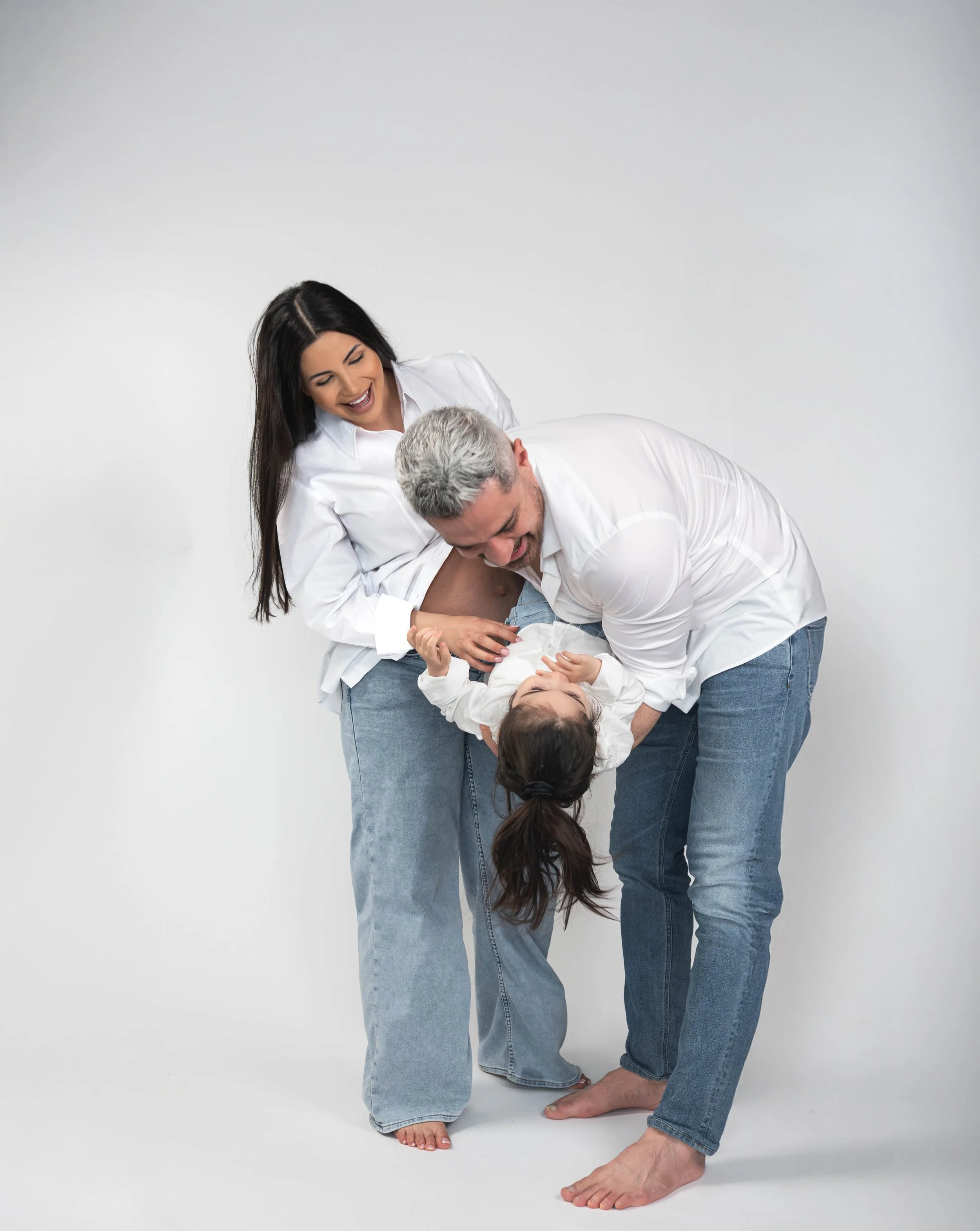 A woman with long dark hair and a man with gray hair and beard, both wearing white shirts and blue jeans, appear to be playfully wrestling or engaging in a lighthearted, humorous struggle against a plain white wall background.