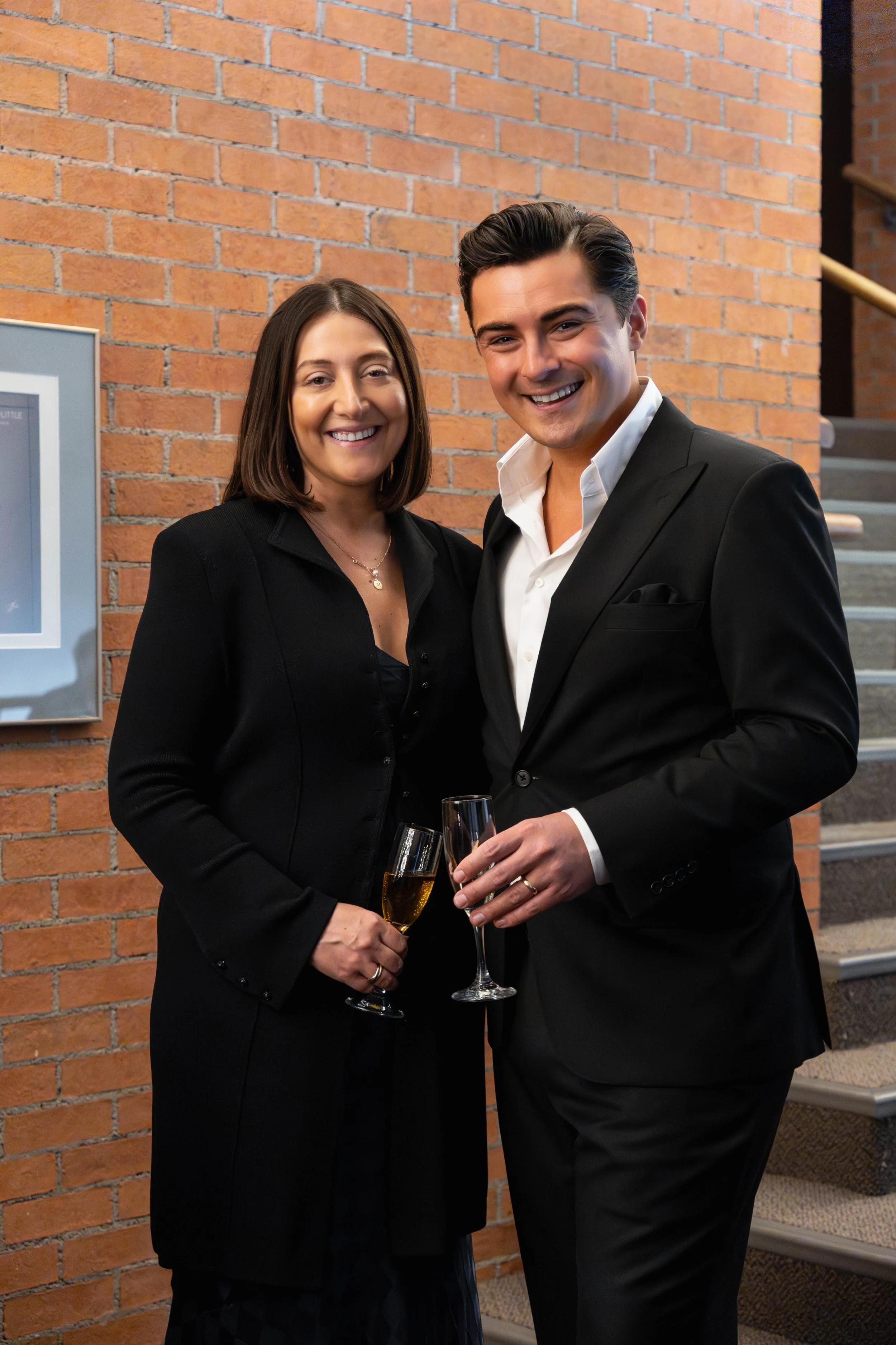 A man and woman dressed in formal attire holding champagne glasses, smiling at an indoor event with a brick wall background.