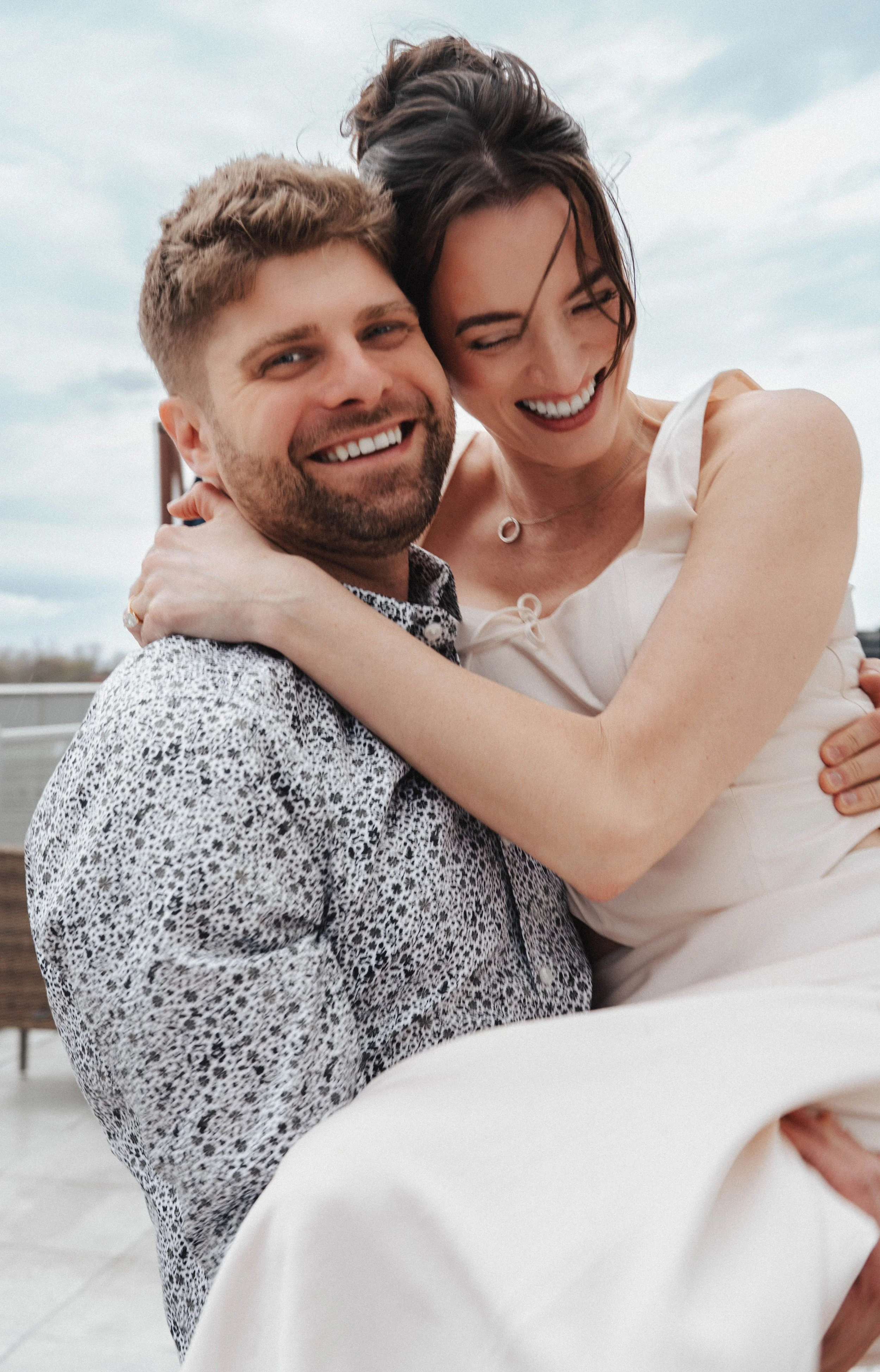 A smiling couple hugging outdoors against a cloudy sky, with the woman in a light-colored dress and the man in a patterned shirt.