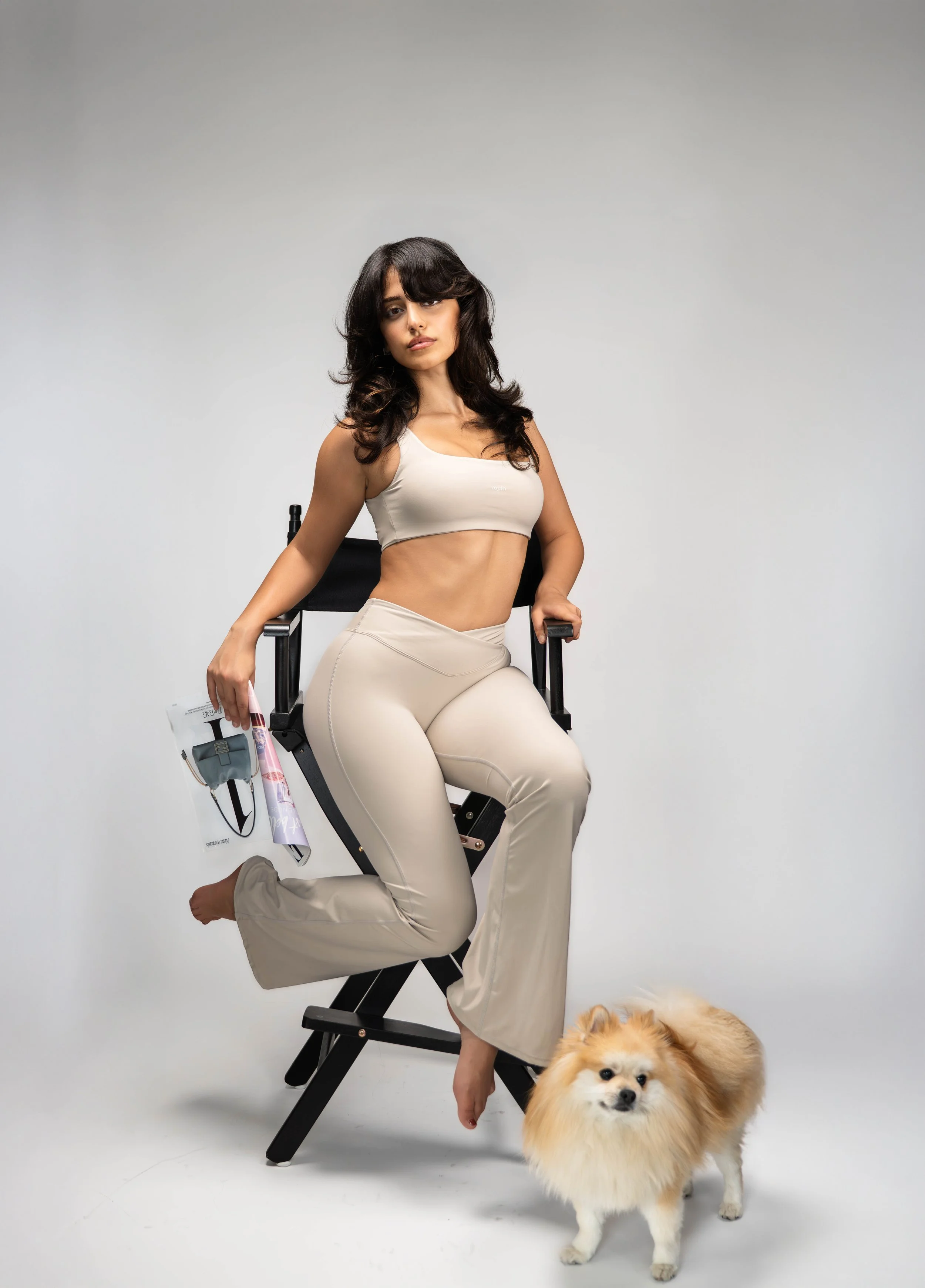 A woman in beige workout clothes sitting on a director's chair, with one leg bent and the other extended. She has long, dark wavy hair and is holding a magazine or newspaper in her right hand. A small, fluffy Pomeranian dog stands next to her on the 
