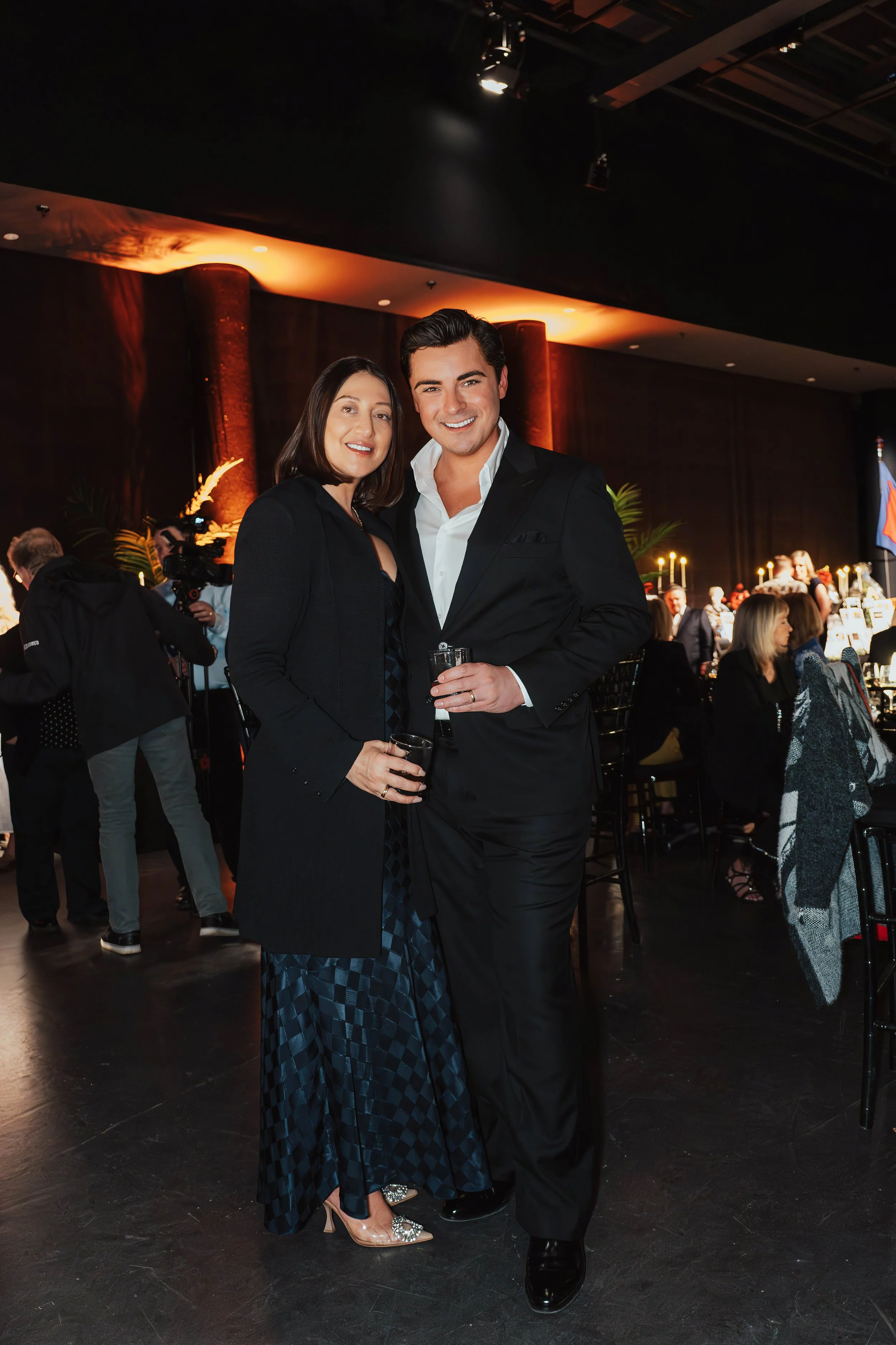 A man and woman dressed in formal attire posing together at an indoor event, holding drinks. The background shows other guests, tables, and dim lighting, indicating a celebration or banquet setting.