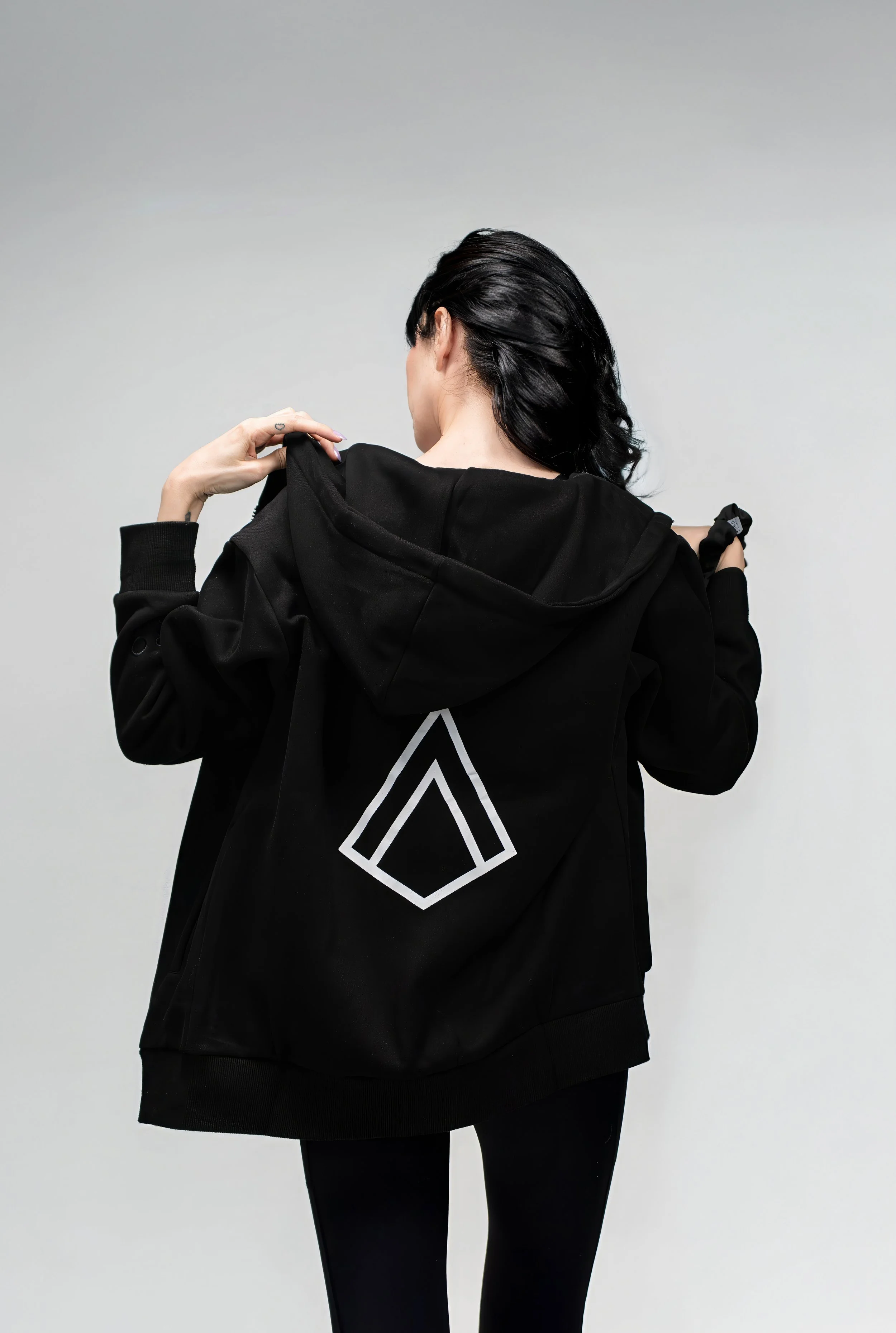 Person with black hair wearing a black hoodie with a geometric logo on the back, facing away from the camera, adjusting the hoodie.